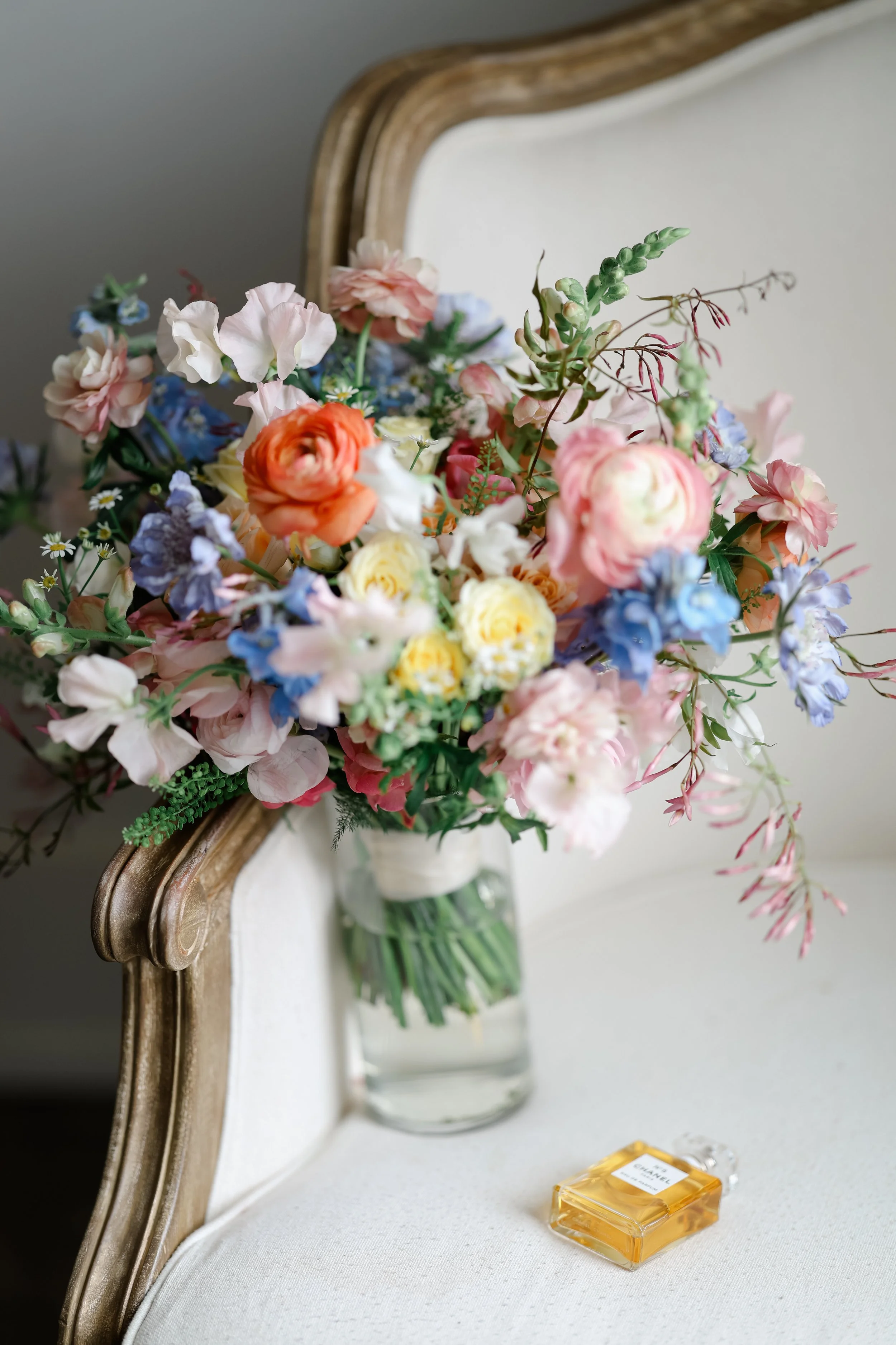 A glass vase with a bouquet of pink, orange, yellow, blue, and purple flowers resting on a cream-colored upholstered piece of furniture with an ornate wooden frame, with a Chanel perfume bottle nearby.