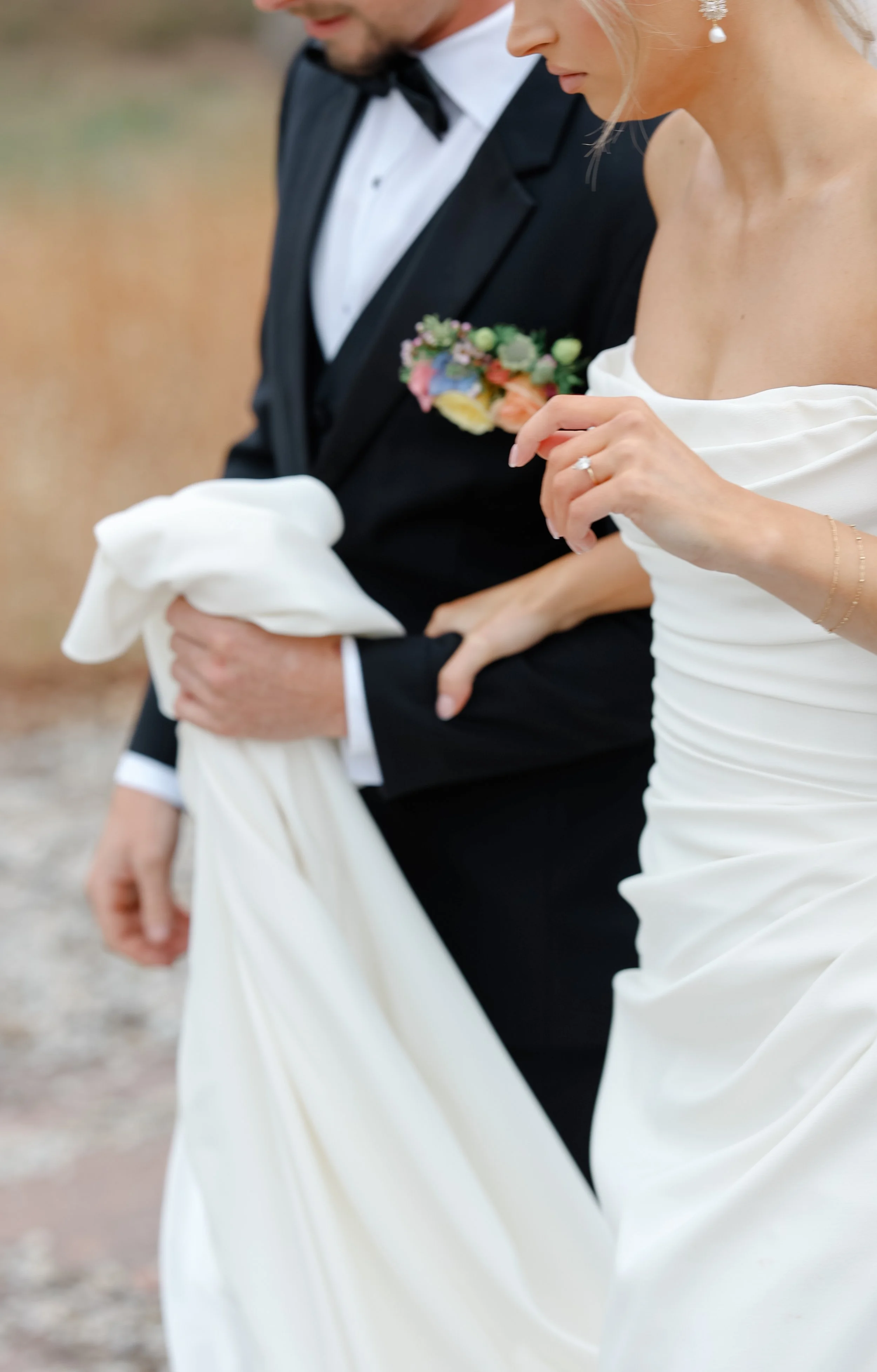 A bride and groom standing outdoors, dressed in formal wedding attire, with the bride holding her wedding dress and showing a diamond ring.