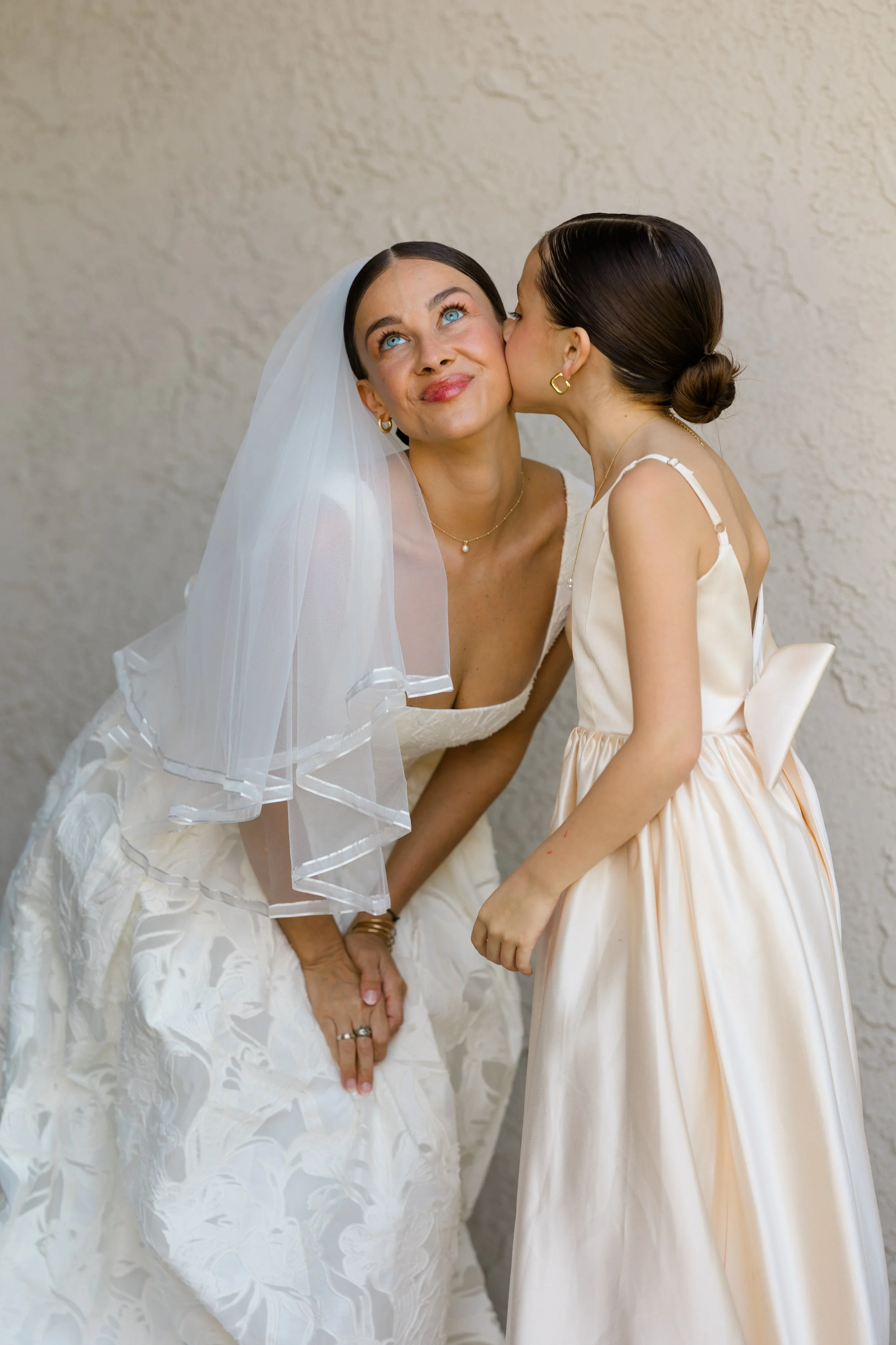 Two women, one in a wedding dress and veil, and the other in a satin dress, sharing a moment of affection, with the woman in the wedding dress smiling and the woman in satin giving a kiss on the cheek, against a plain textured wall.