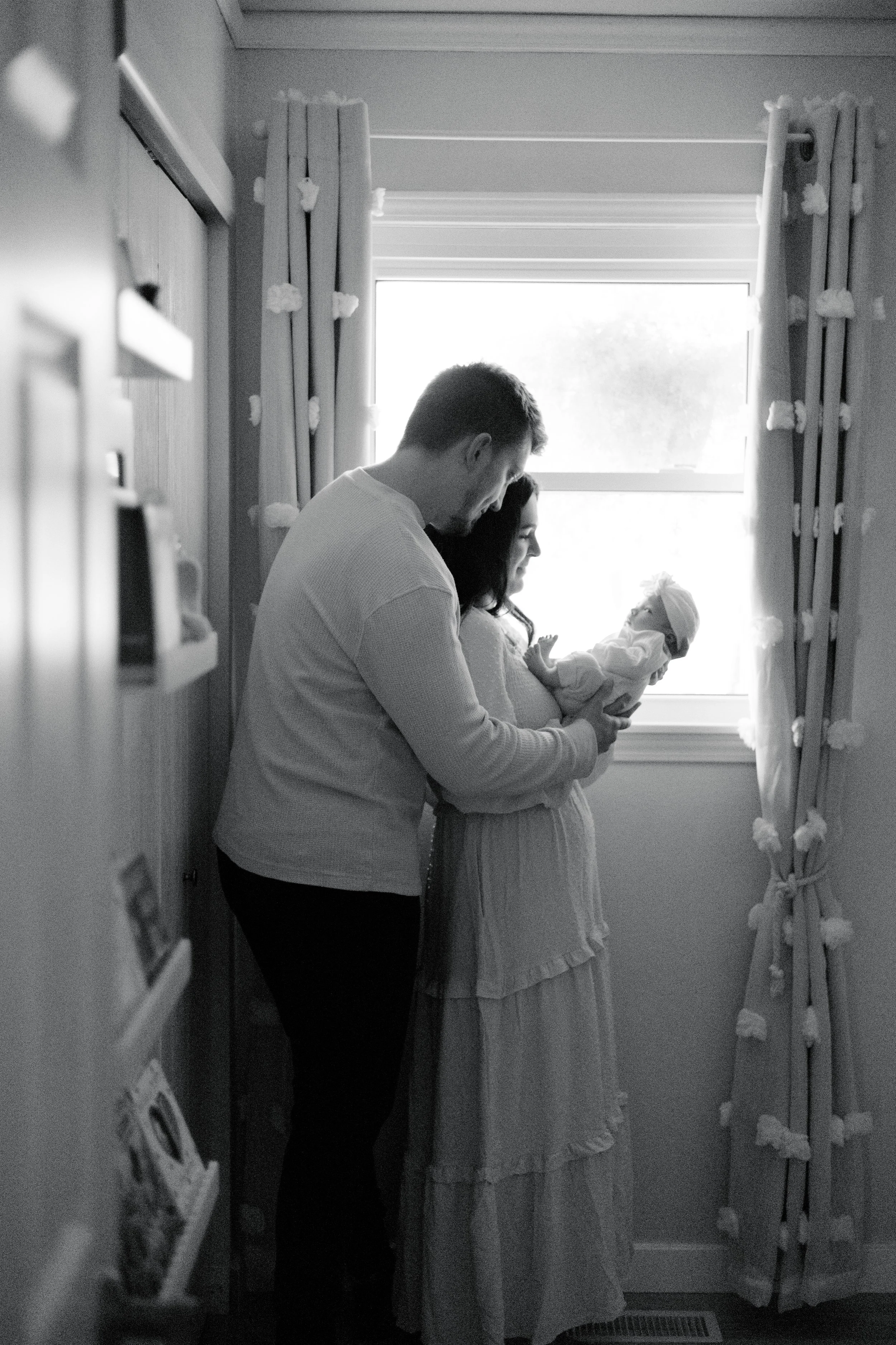 A man and a woman standing near a window in a hospital room, holding a newborn baby girl. The woman is dressed in a hospital gown, and the man is in casual clothes. The room has curtains and a window with sunlight coming in.