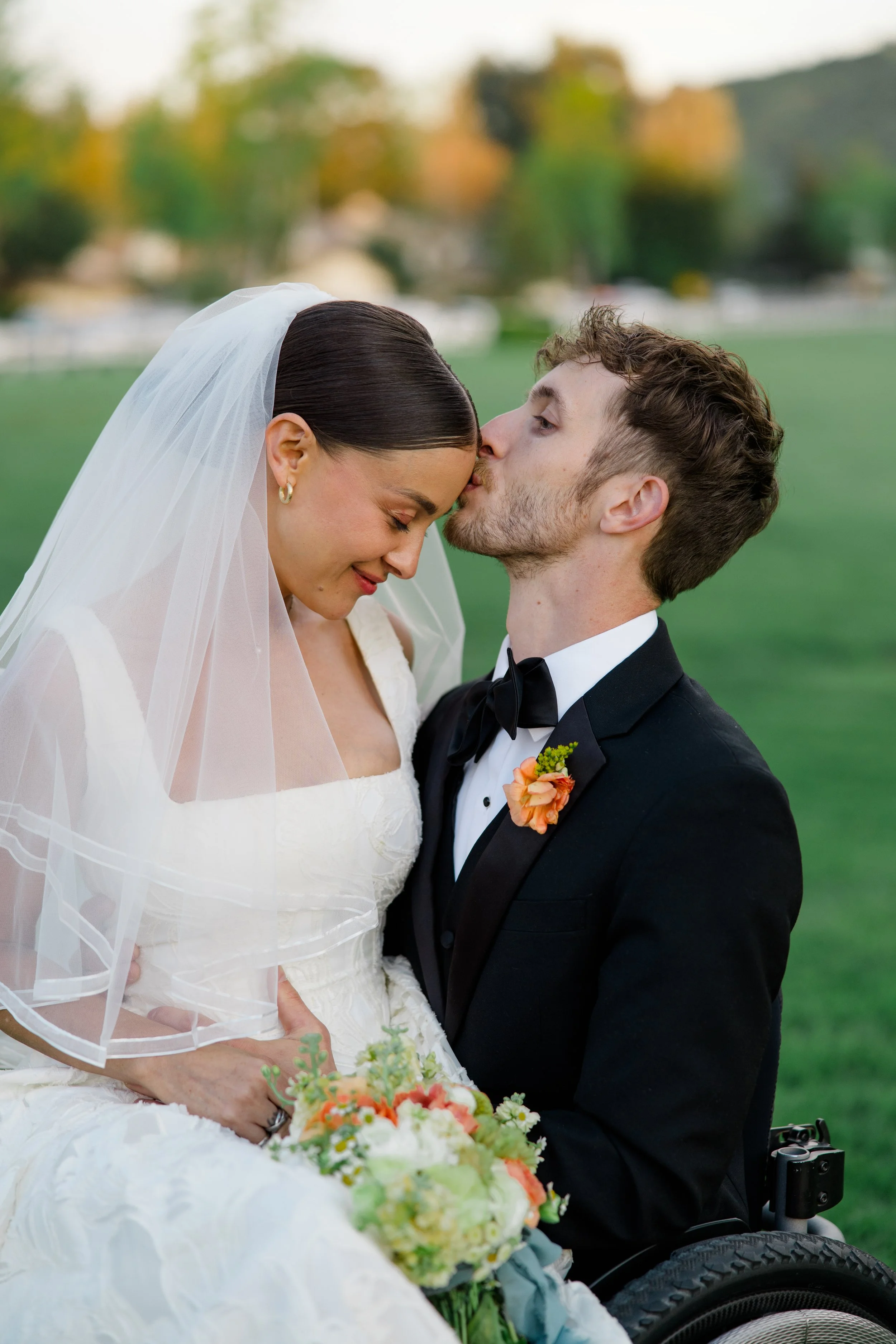 A bride and groom sharing a kiss outdoors, the bride holding a bouquet of flowers, and the groom in a wheelchair, with a blurred background of trees and a field.