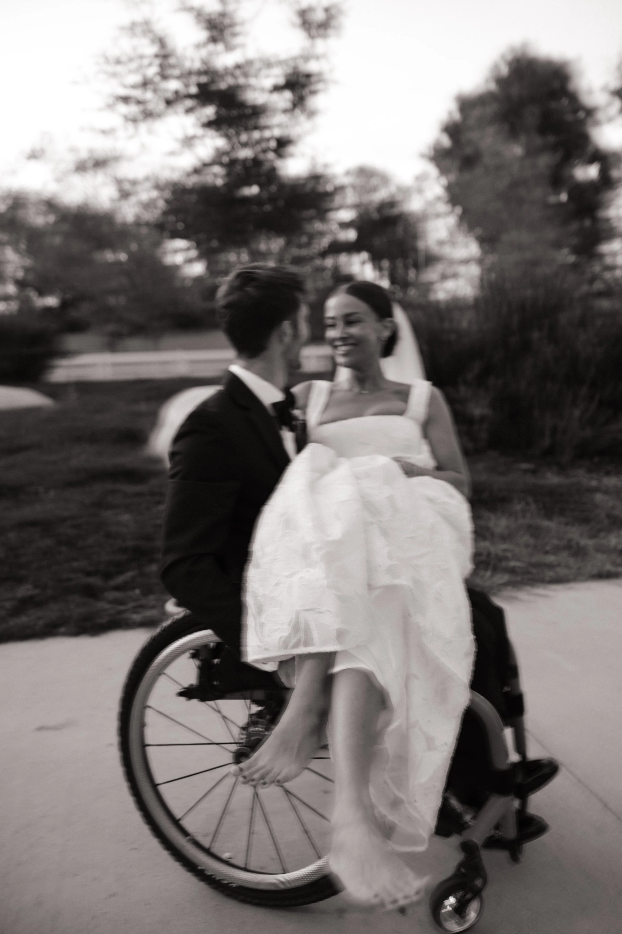 Black and white photo of a groom in a suit and a bride in a wedding dress, smiling at each other. The bride is in a wheelchair outdoors, with trees in the background, suggesting a park or garden setting.