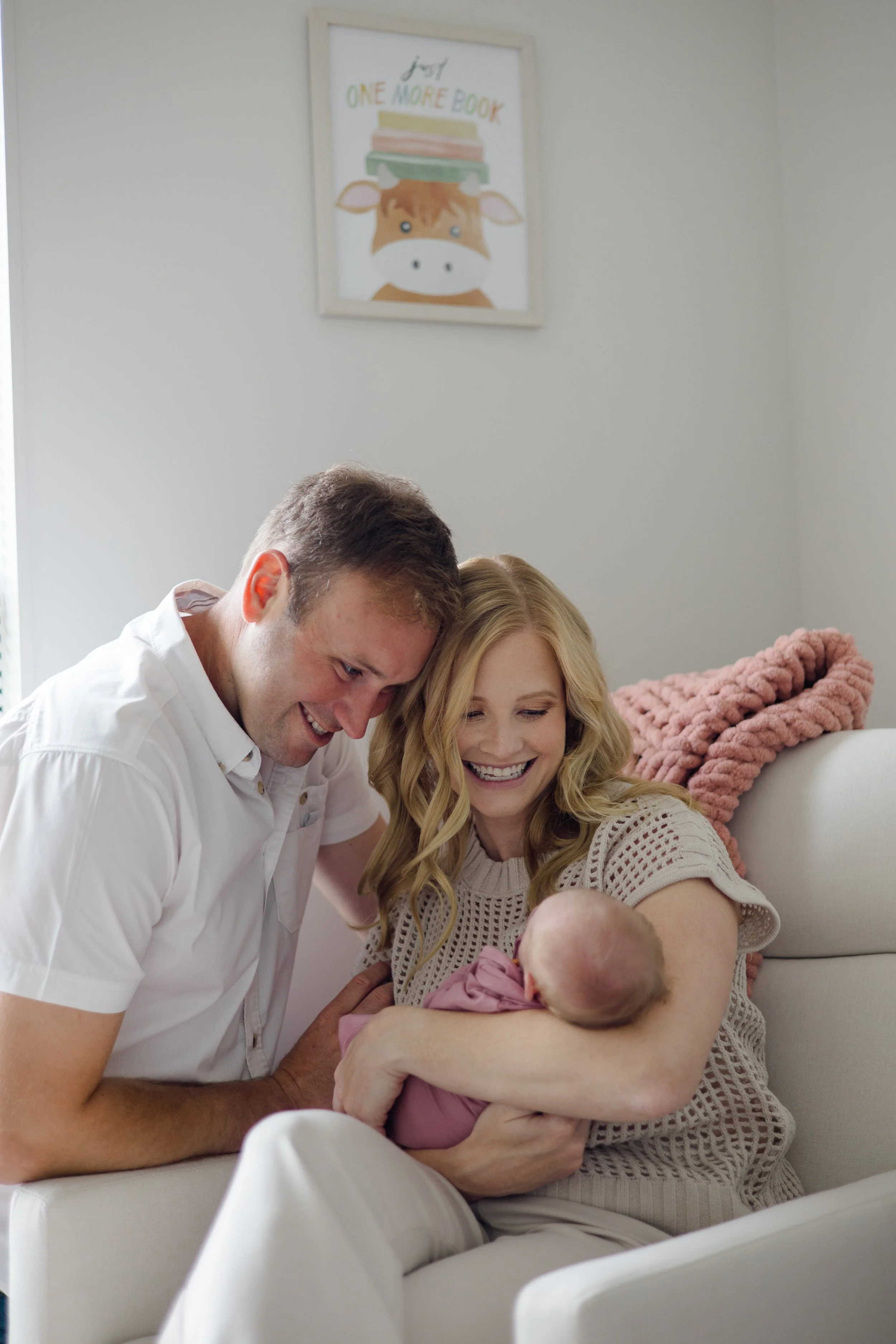 A man and a woman with light skin and blonde hair hold a newborn baby while smiling, sitting on a light-colored sofa in a cozy room with a pink chunky knit blanket on the armrest. There is framed wall art with a cartoon cow and the text 'just one mor