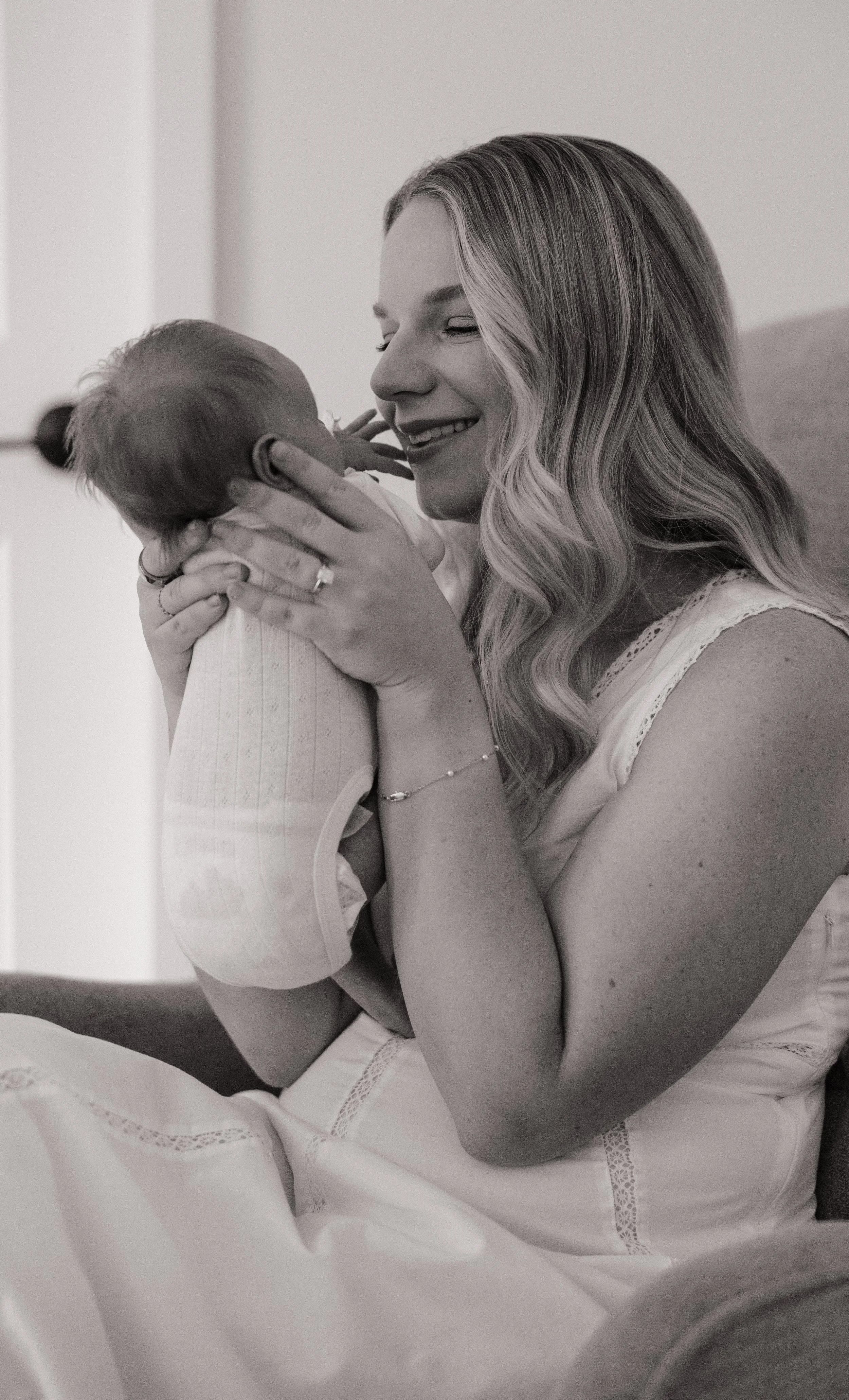 Black and white photo of a woman holding a baby close, smiling and looking at the baby, with soft background and natural light.