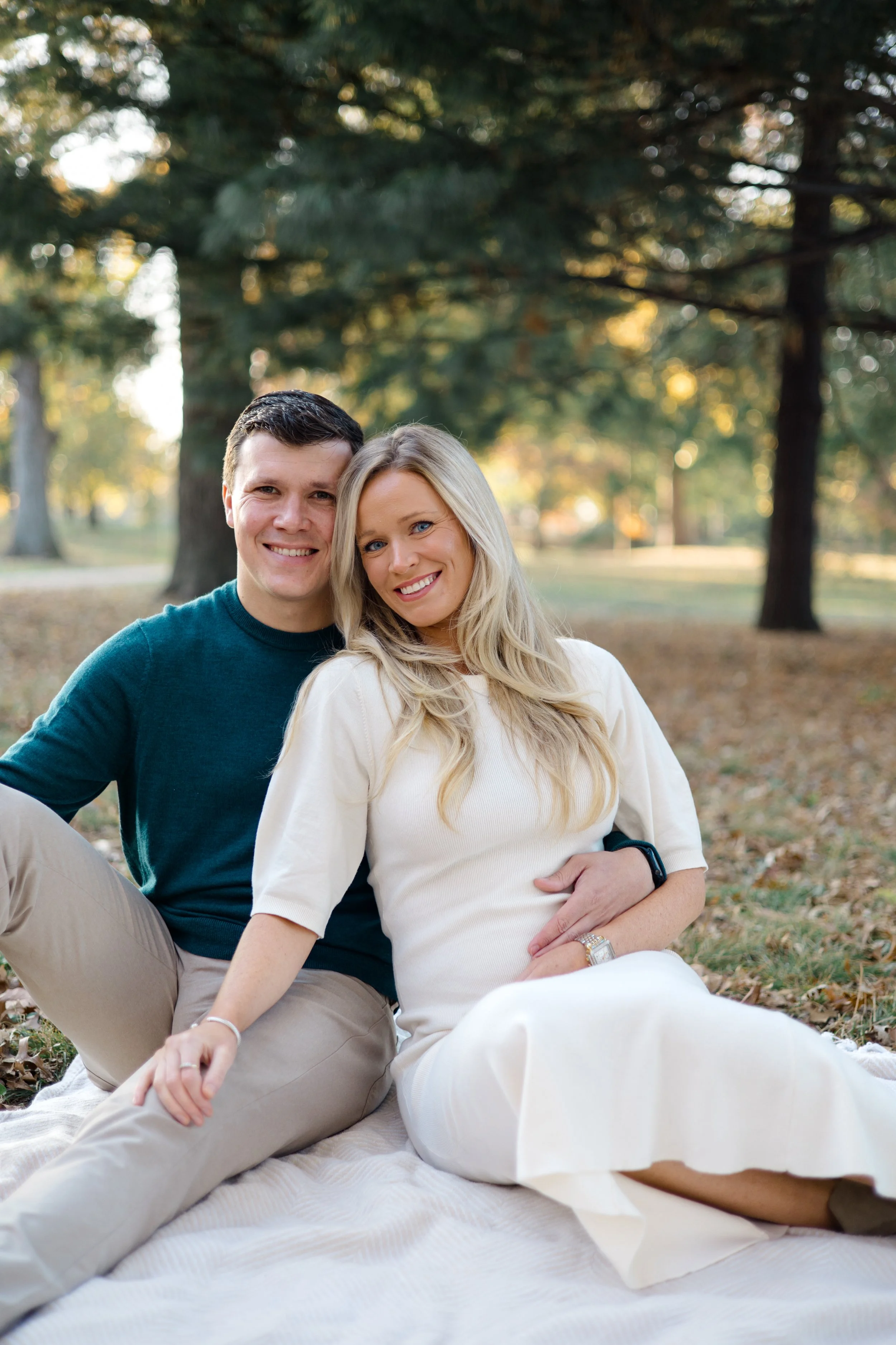 A couple sitting on a blanket on the grass in a park with trees in the background. The man has short dark hair and is wearing a teal shirt. The woman has long blonde hair and is wearing a cream-colored dress.