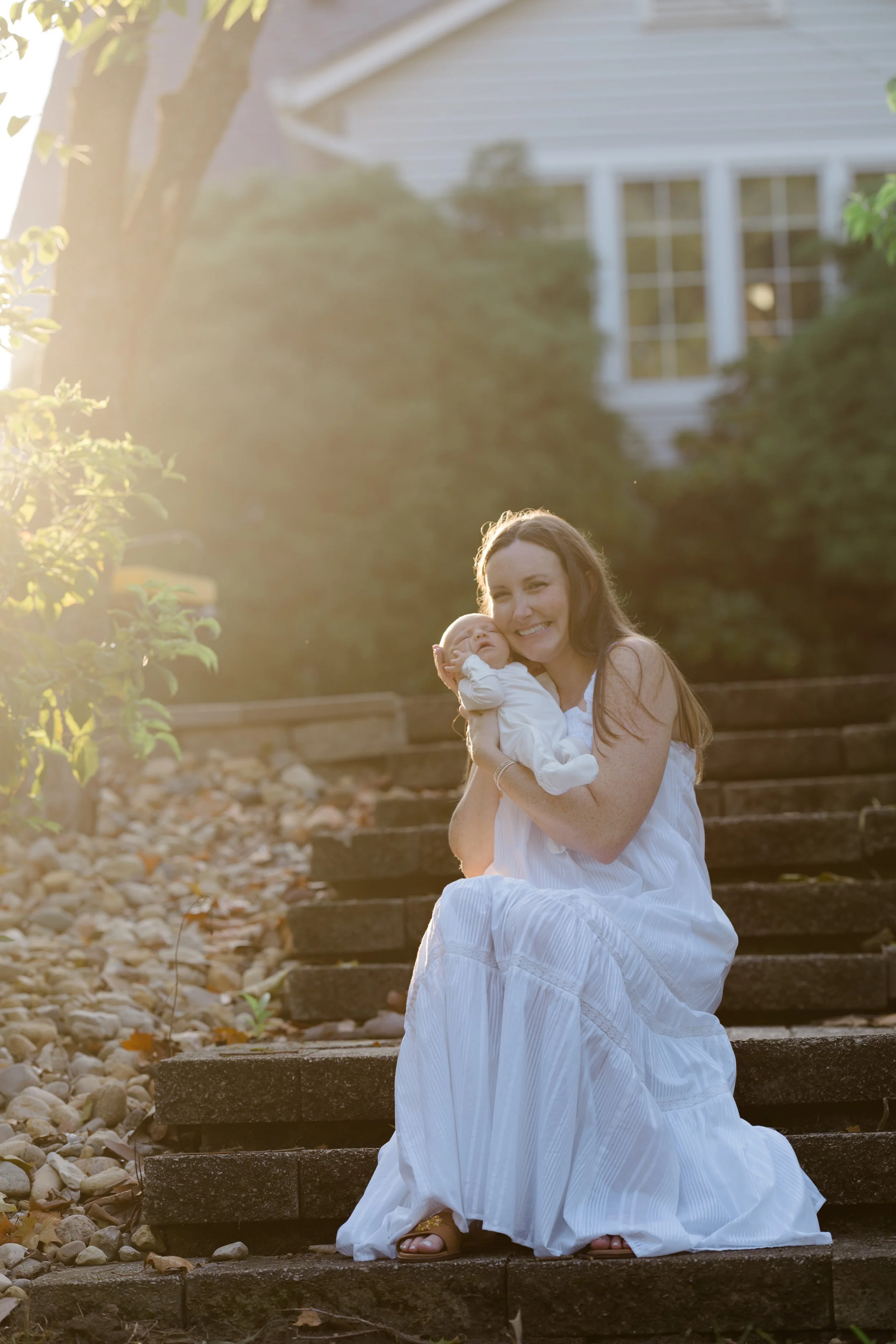 A woman in a white dress sitting on outdoor steps, holding a baby, with sunlit trees and a house in the background.