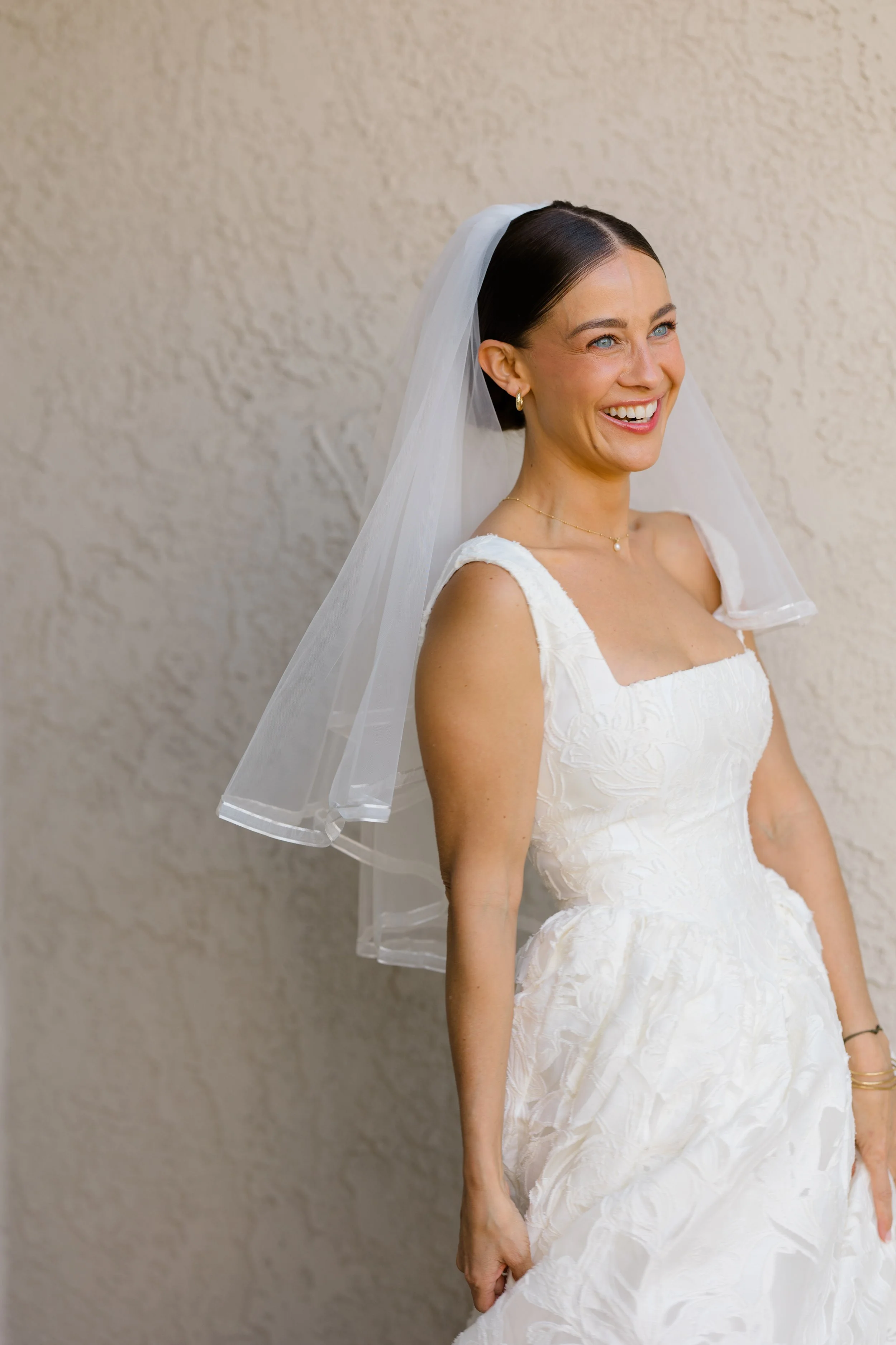 A woman in a wedding dress with a veil, smiling, standing by a beige textured wall.