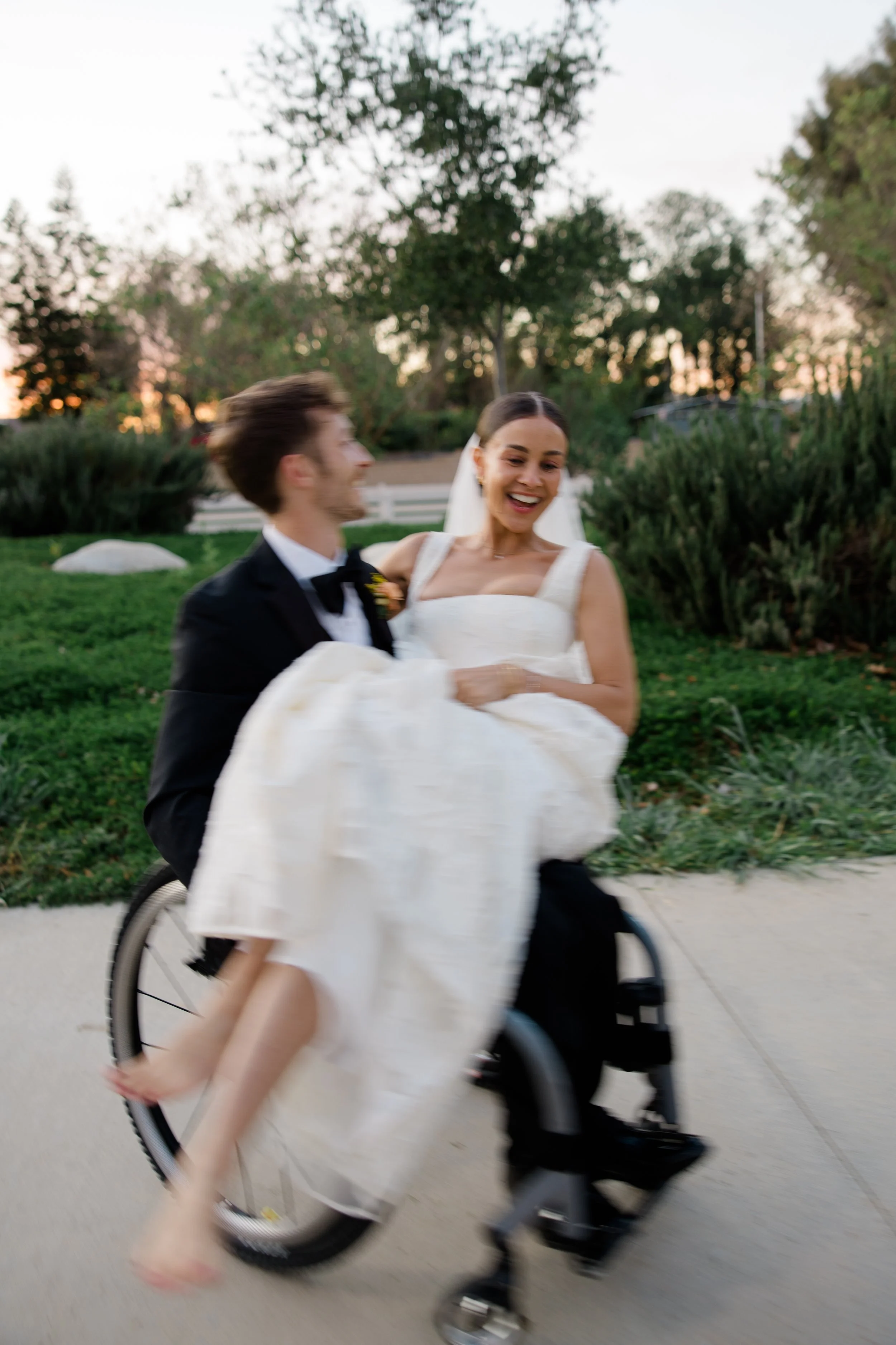 A bride in a white wedding dress smiling while riding a bicycle with a groom in a tuxedo, outdoors near trees during sunset.