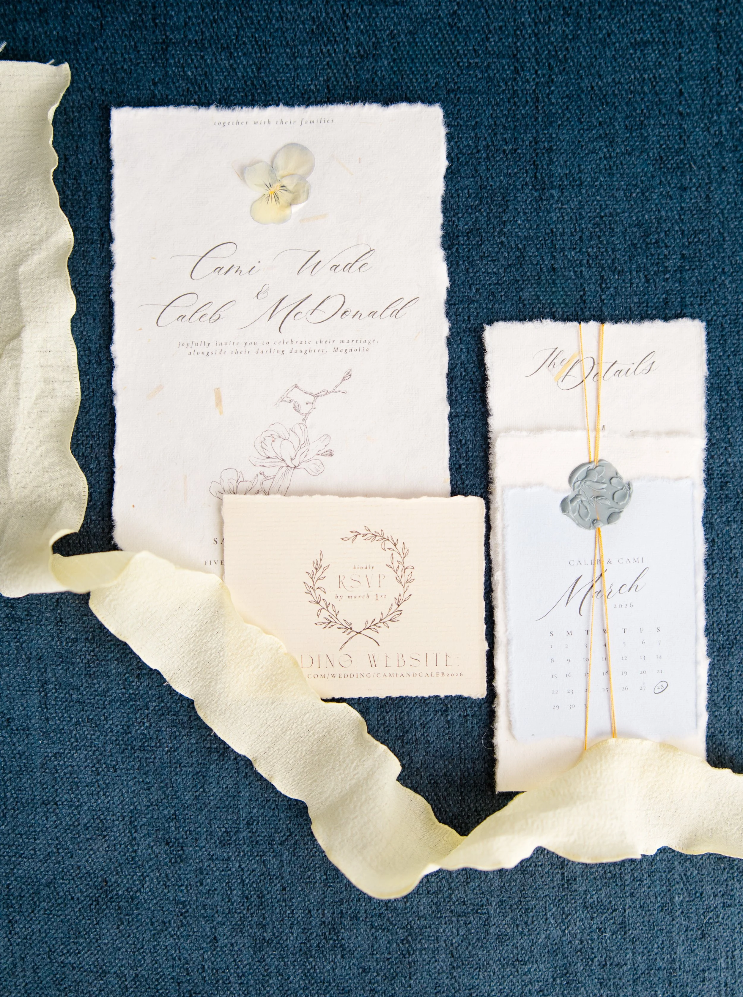 Collection of wedding invitations, RSVP card, and a gray floral ring with yellow string on a dark blue textured surface, with a beige torn paper ribbon overlay.