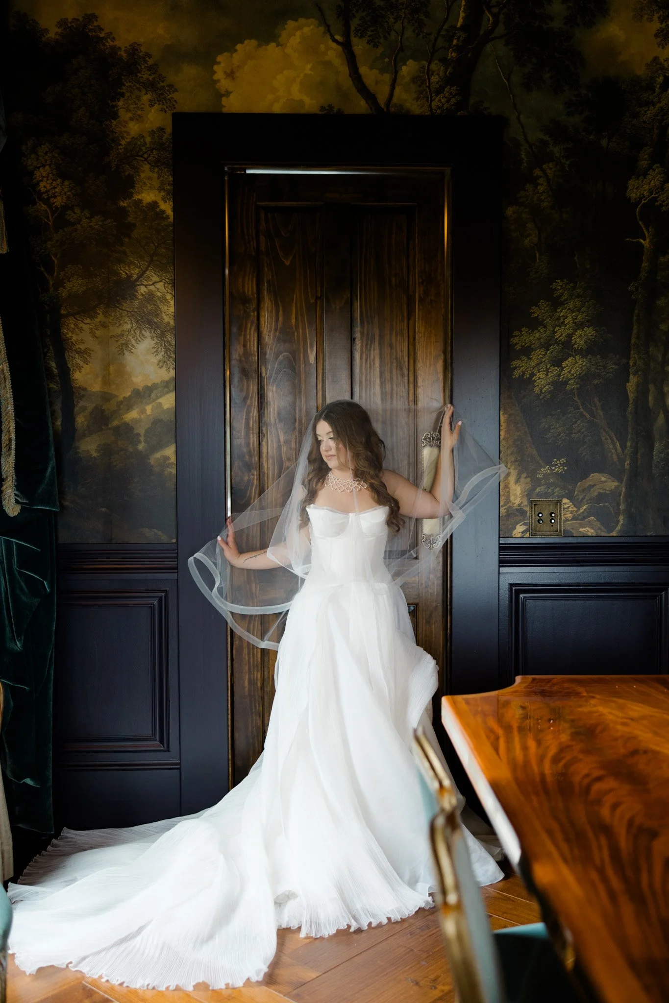 A woman in a white wedding dress and veil standing in front of a wooden door, holding the veil with both hands, with a nature-themed mural in the background.