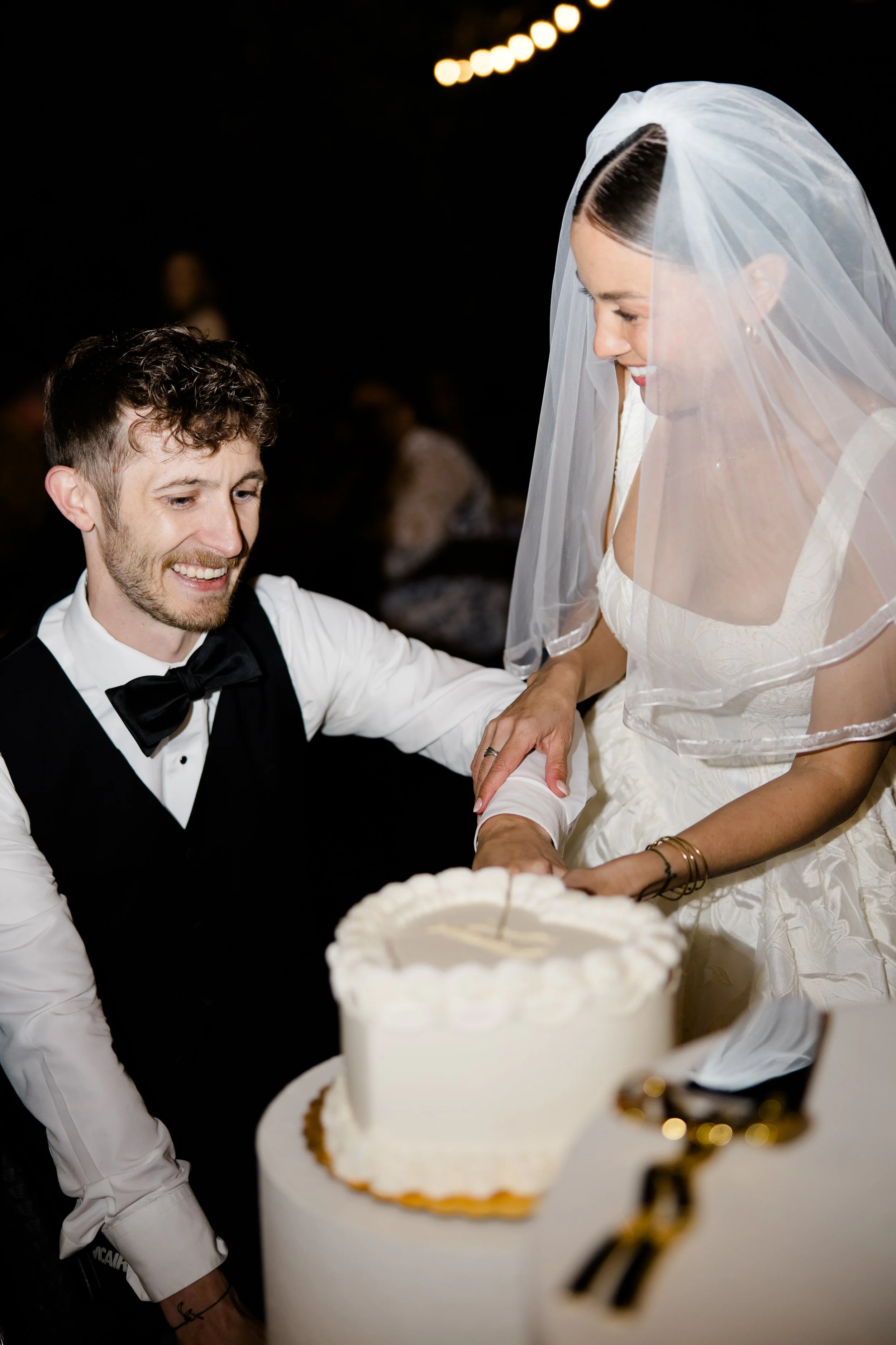 A bride and groom smiling at each other during their wedding reception, with a wedding cake in the foreground.