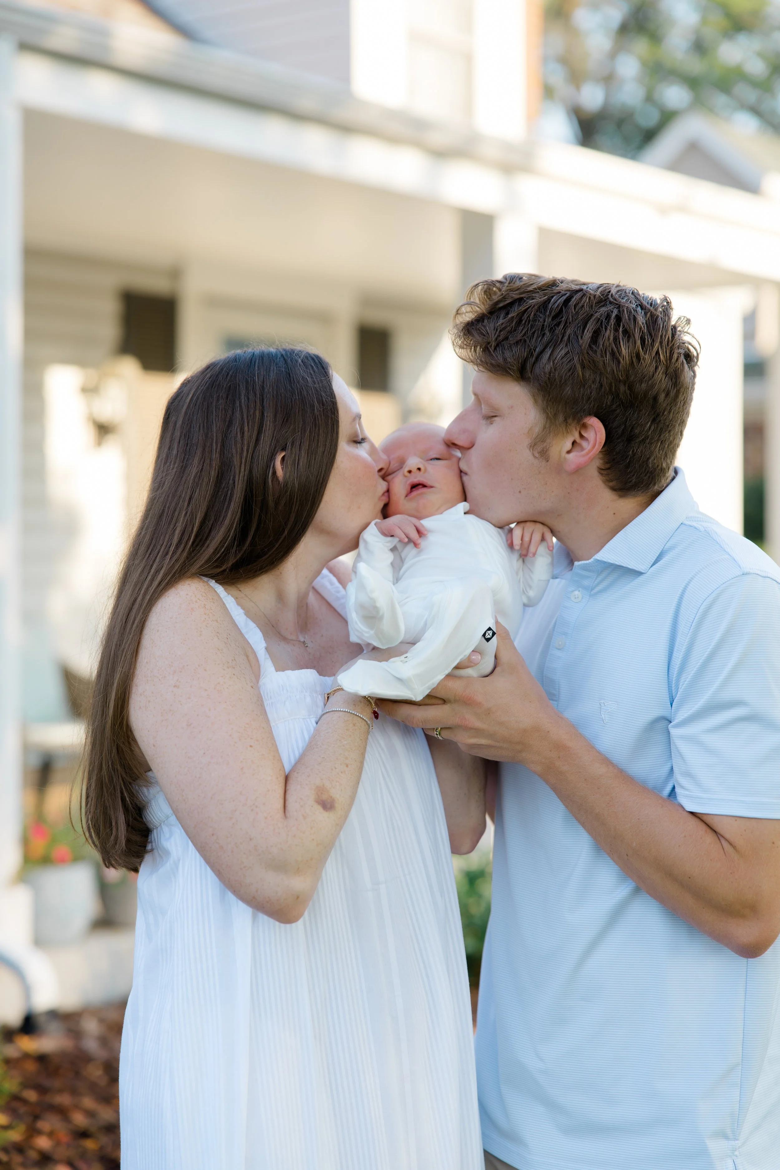 A family of three, a woman, a man, and a baby, sharing a kiss outside on a sunny day, with a house in the background.