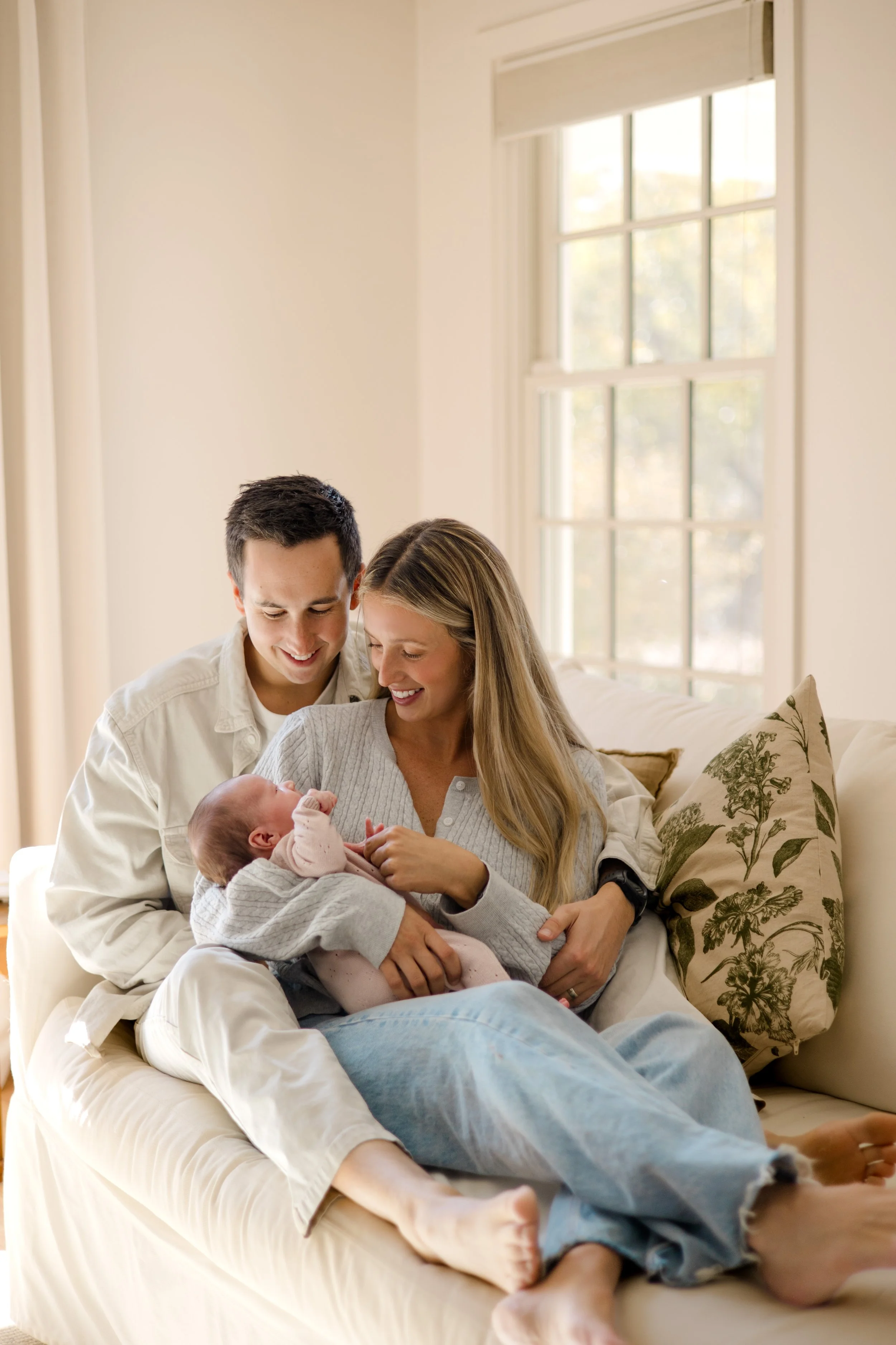 A family of four, including two adults and two children, sitting together on a sofa in a well-lit room with a large window, celebrating the arrival of a newborn baby.