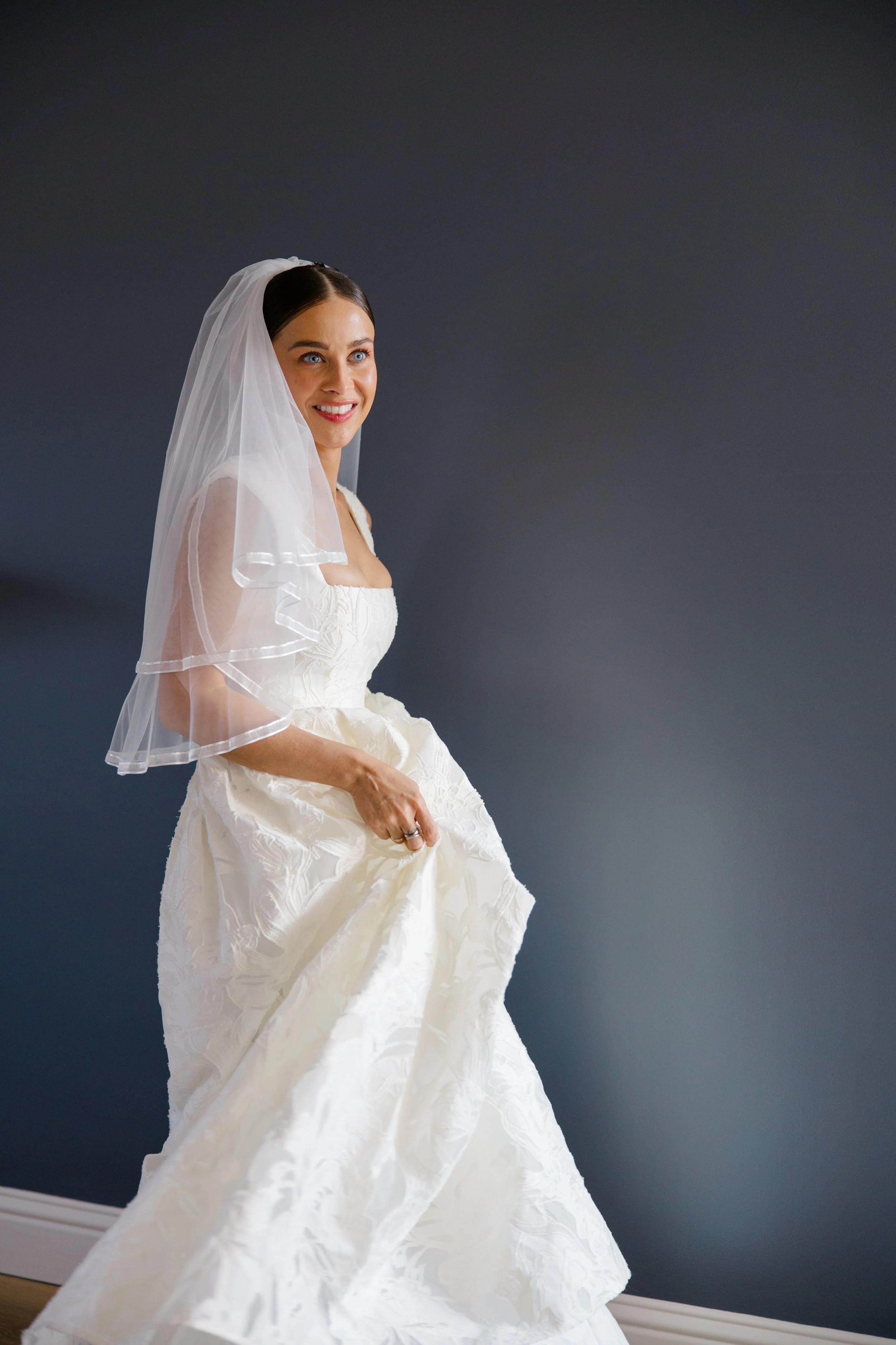 A woman in a wedding dress holding the skirt of her gown, standing against a dark gray wall, smiling softly.