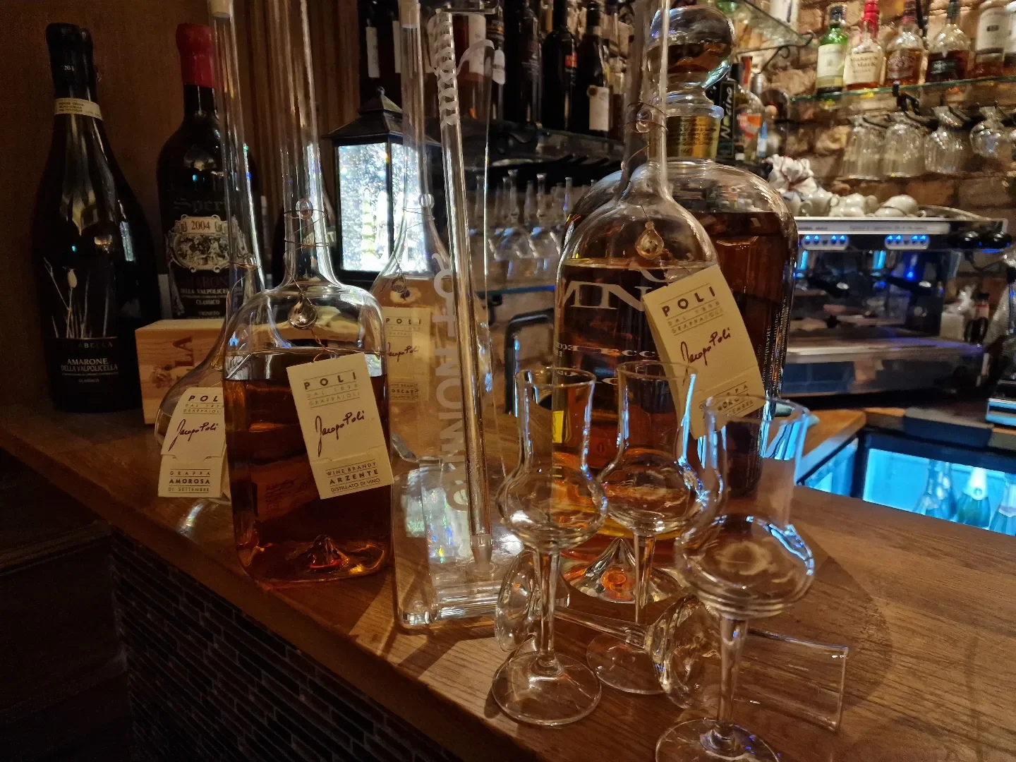 Bild av en bar med flera flaskor av sprit och vin, inklusive en flaska med etiketten 'Japo Pali'. Det finns även ett glas med en liten mängd sprit och andra tomma glas på en träbänk.