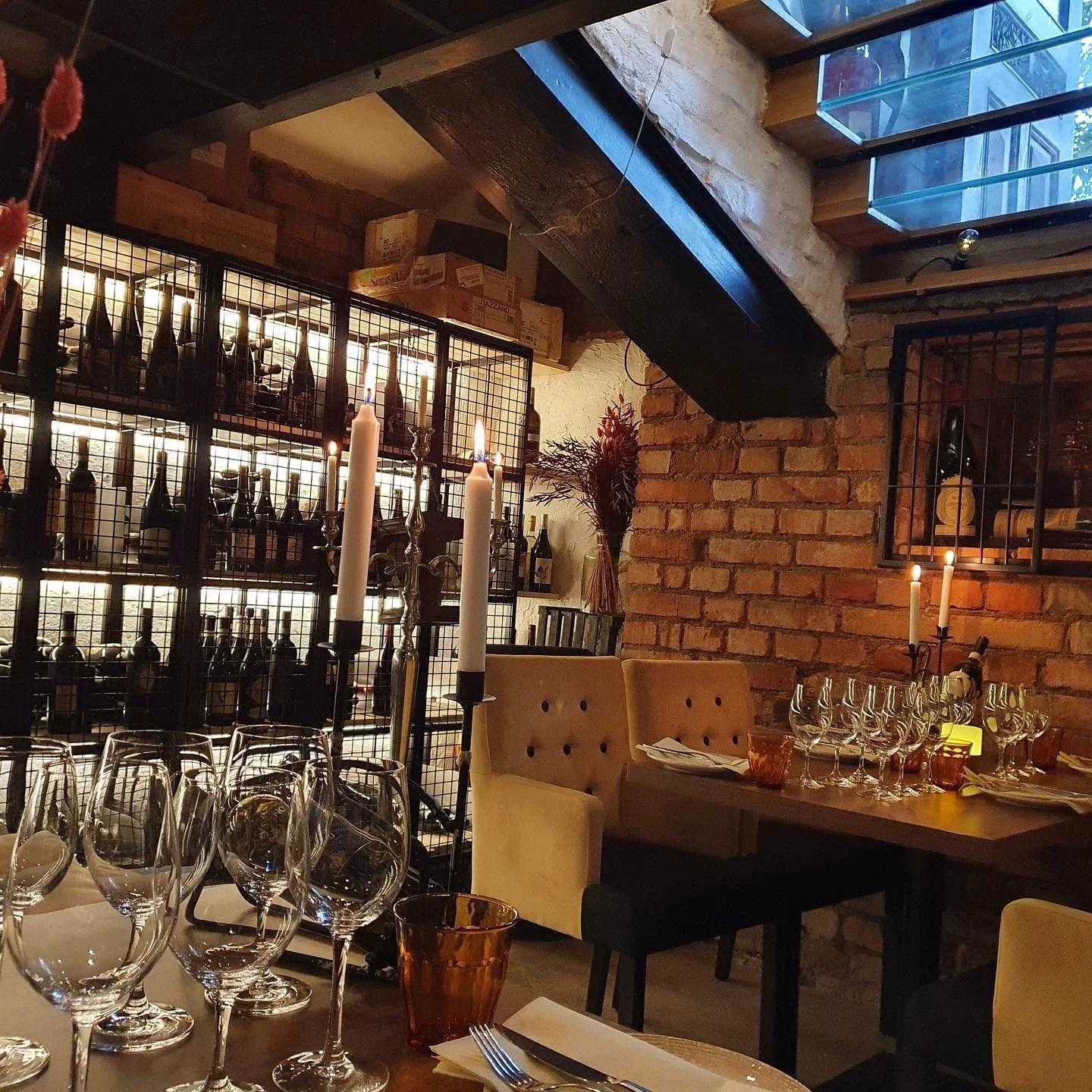 Inomhusbild av en restaurang eller matsal med vinflaskor på hyllor, flera tomma vinglas, tända ljus och brickor för att duka, väggar av tegel och ett tak av trä, ett fönster med glas som ger naturligt ljus.