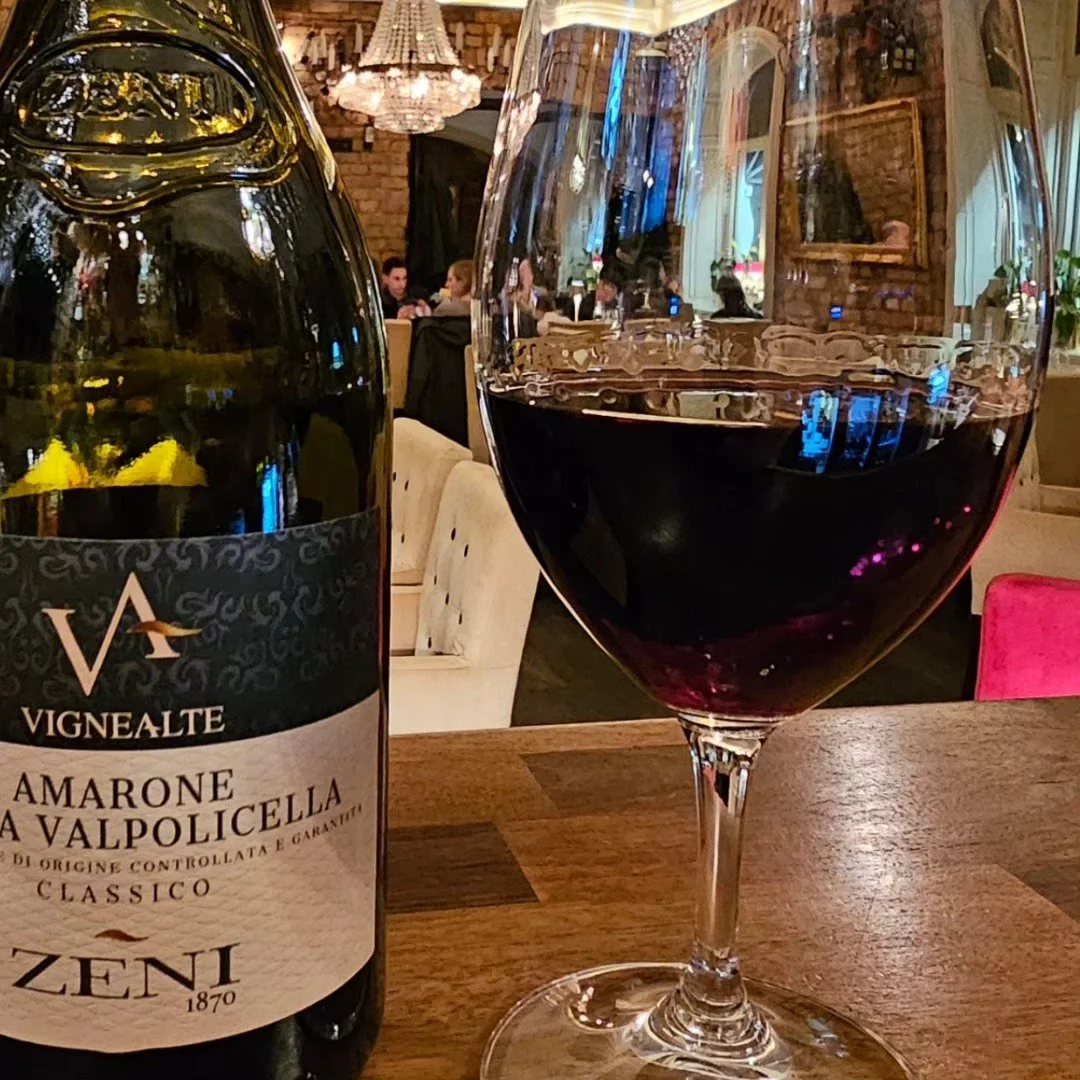 Ett glas rött vin och en vinflaska på ett bord i en restaurang, med människor som äter och umgås i bakgrunden.