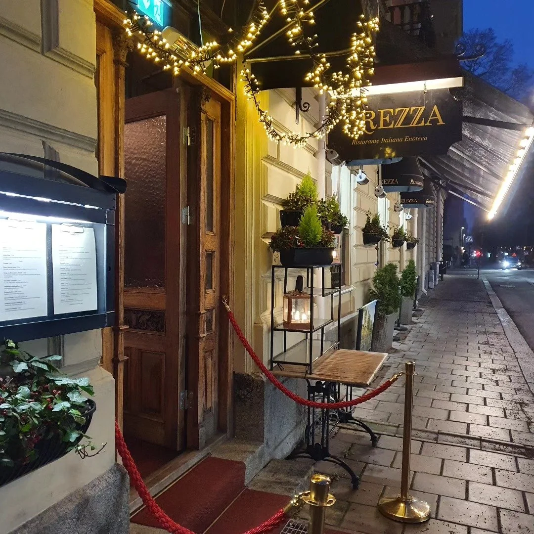 Ingång till en italiensk restaurang med namnet Rezza, dekorerad med ljusslingor och växter utanför, på en kväll.