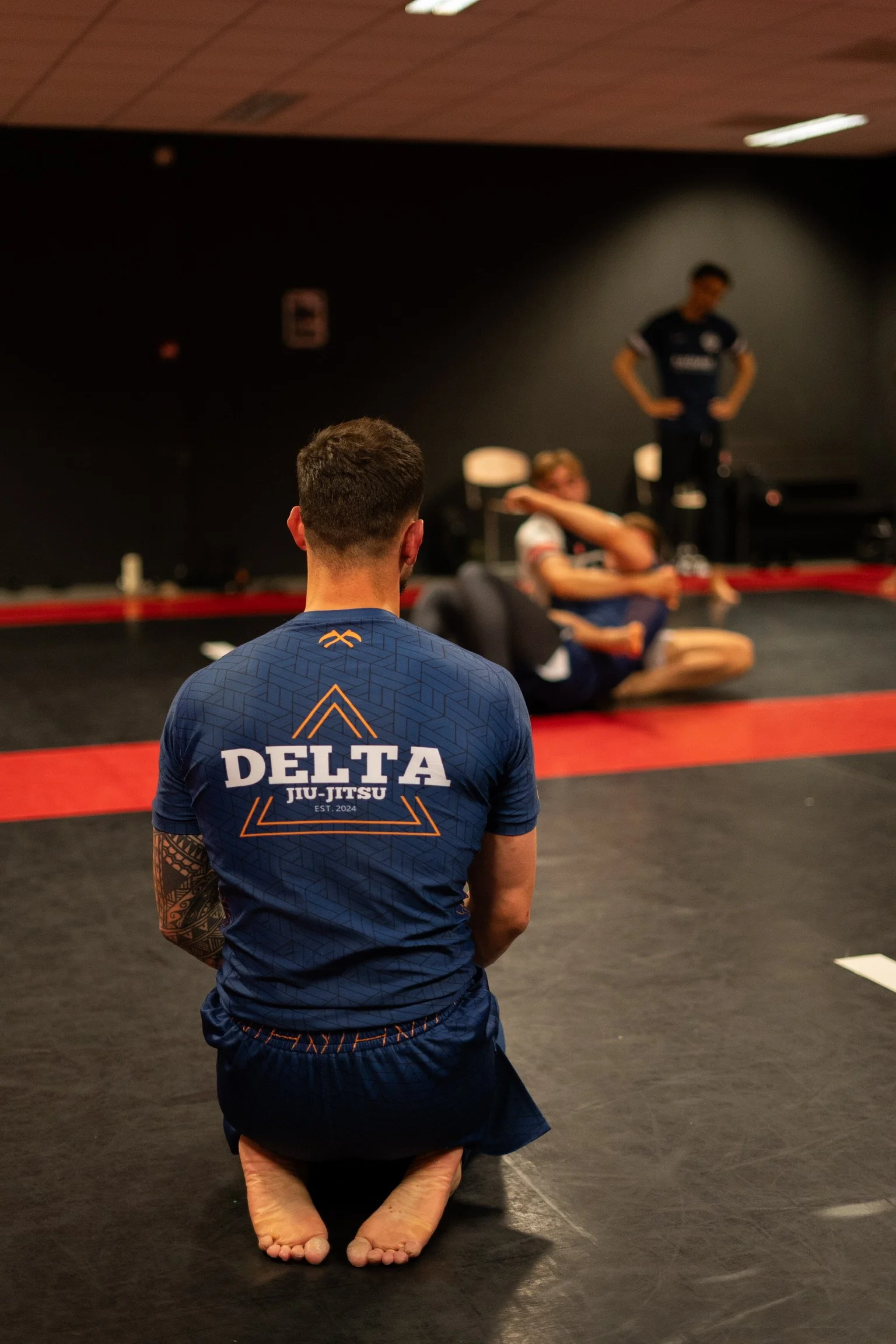 Een persoon in een Delta Jiu-Jitsu shirt zit met gekruiste benen op de grond in een sportschool, terwijl anderen in de achtergrond voor training of ontspanning zitten.