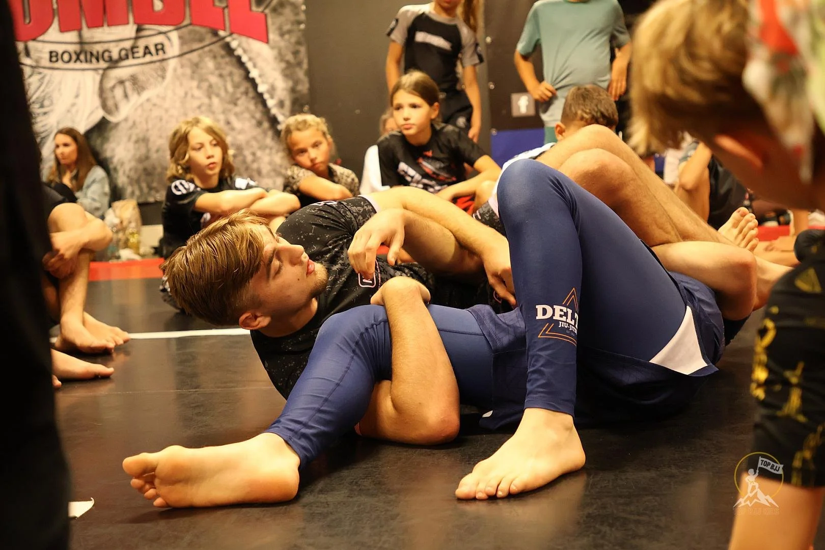 Mensen die bjj beoefenen, liggen op de mat en trainen, terwijl anderen toeschouwers in de achtergrond zitten en kijken.