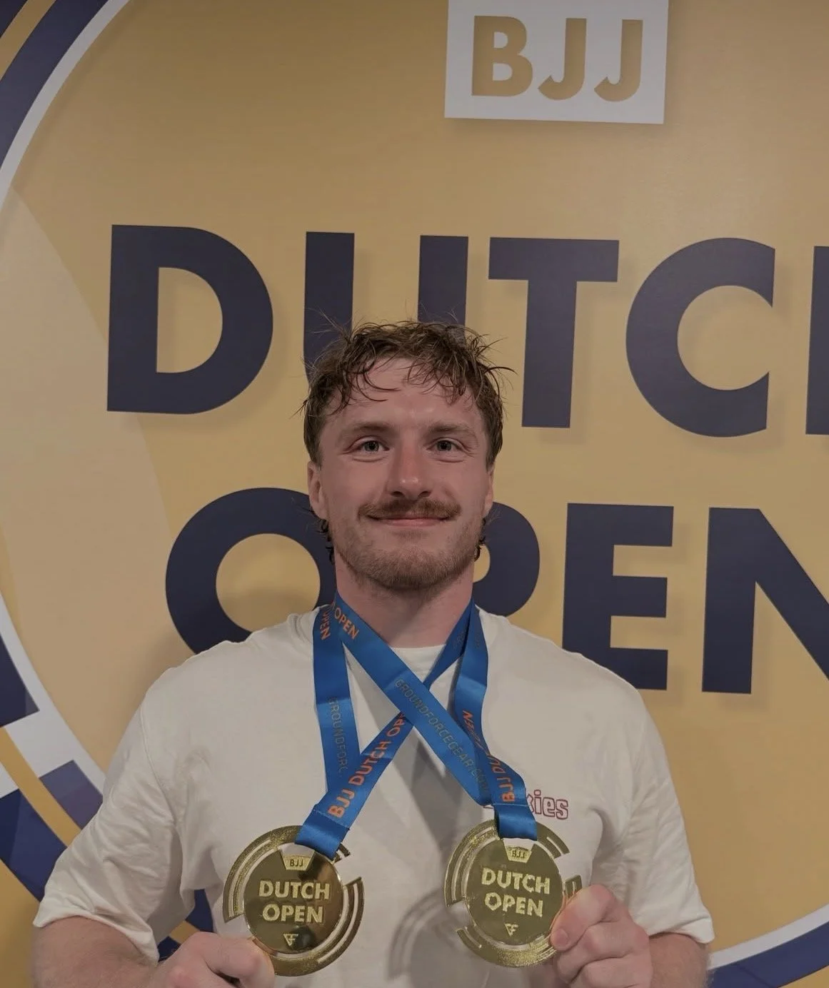 Man met korte, vochtige haar en een baard, die twee gouden medailles vasthoudt met de tekst "Dutch Open" erop, terwijl hij voor een banner staat met de woorden "BJJ Dutch Open" in de achtergrond.