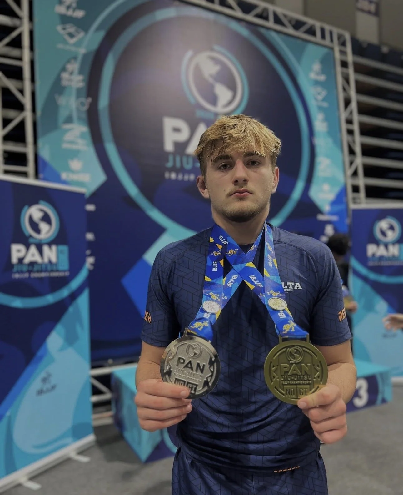 Een jonge man in een blauw sportshirt met gouden en zilveren medailles, poseert op een podium met een achtergrond van banners met het PAN Jiu-Jitsu logo, en toont zijn medailles.