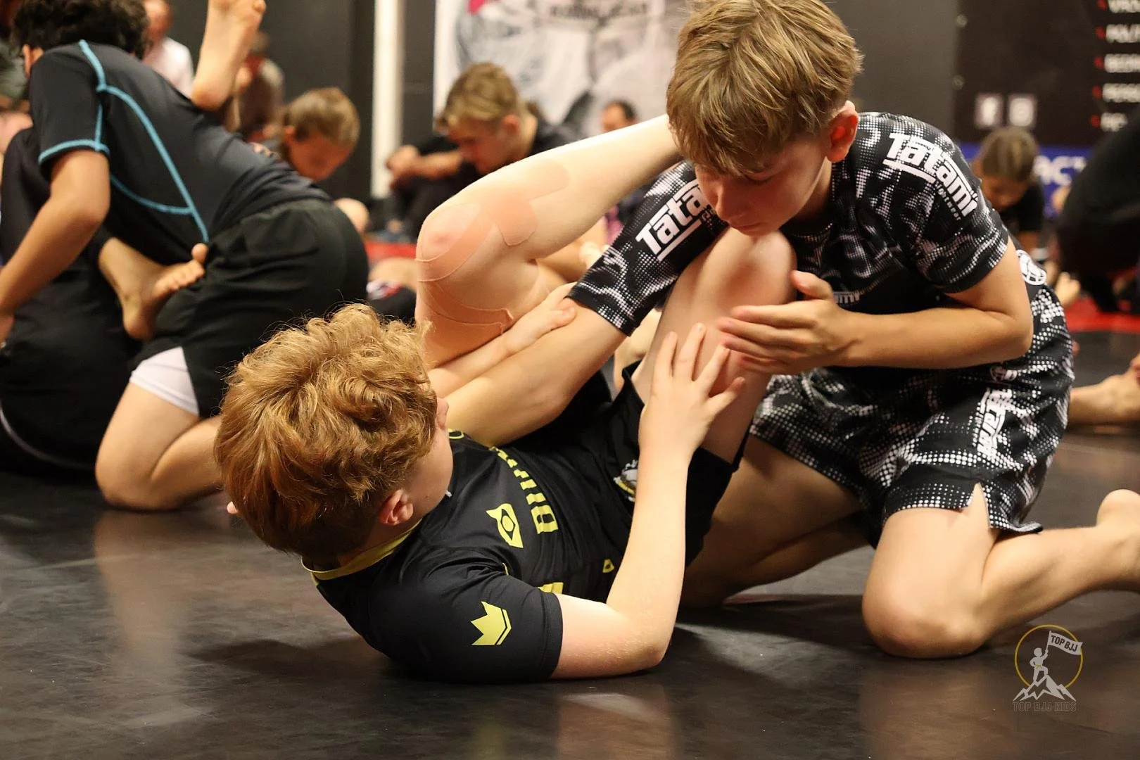 Twee jonge jongens oefenen Braziliaans jiu-jitsu op de mat, met meerdere andere mensen op de achtergrond.