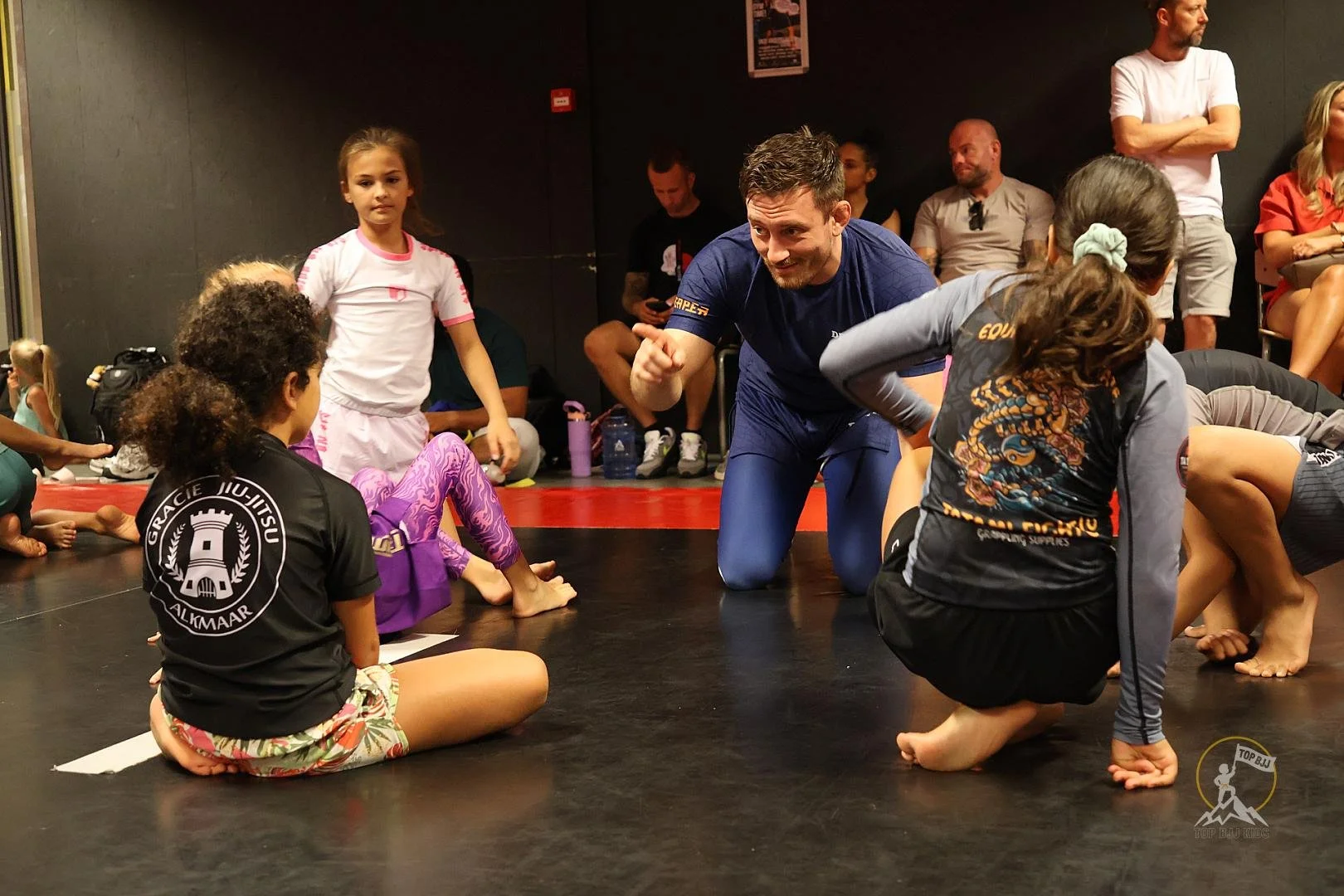 Groep kinderen en een instructeur bij een jiu-jitsu training, met enkele toeschouwers op de achtergrond, binnen een sportschool.