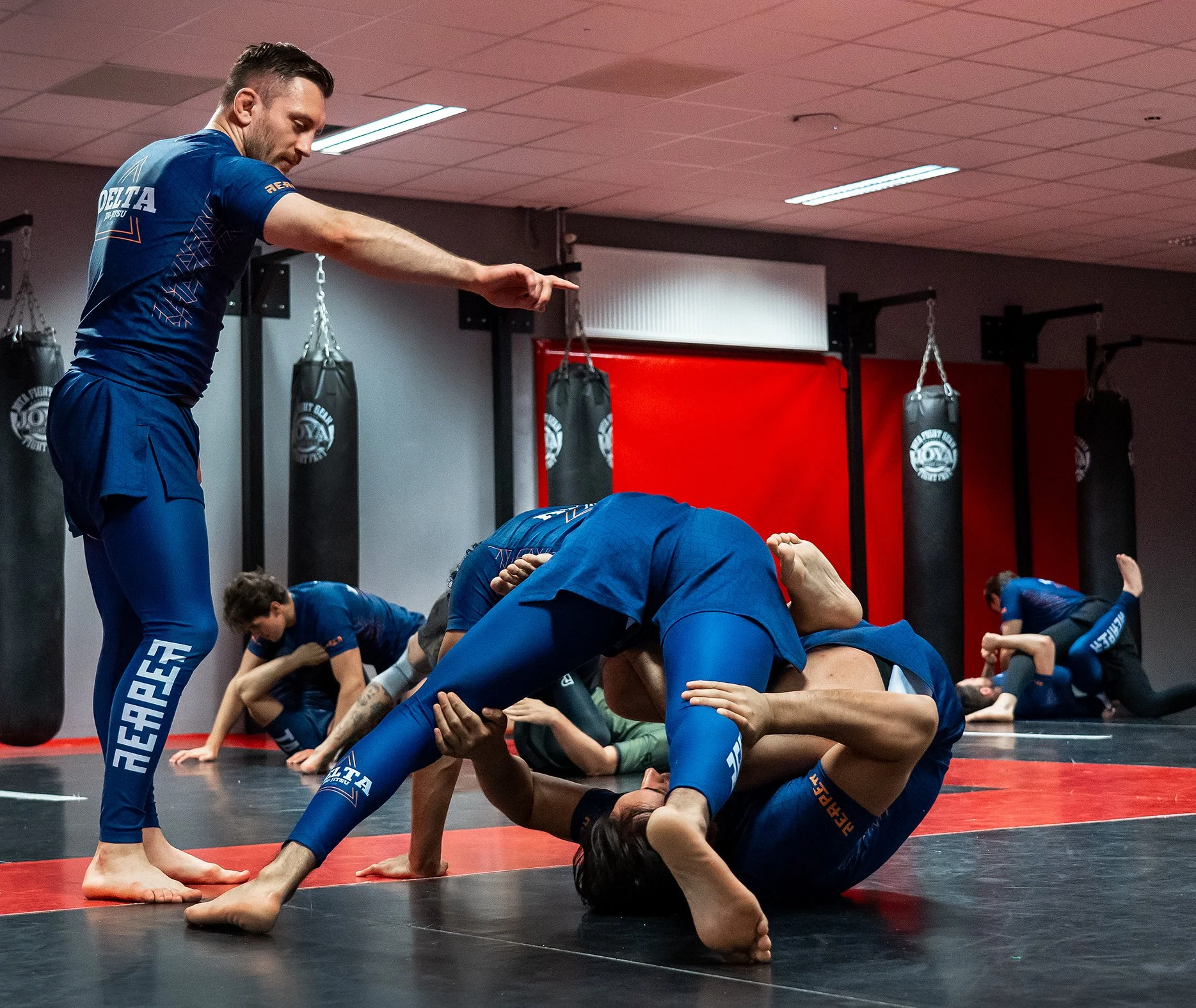 Mannelijke en vrouwelijke jongeren trainen in een sportzaal met matten en bokszakken, oefenen een groepstraining. Een man begeleidt en coacht de groep.