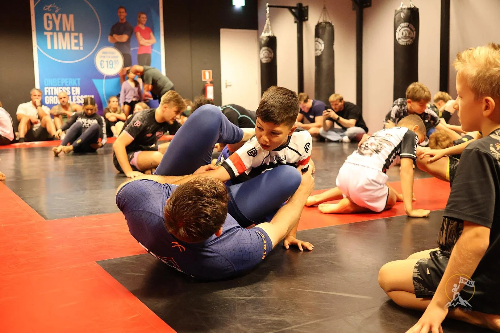 Kinderen die aan Brazilian Jiu-Jitsu training doen op een mat, met een instructeur die hen helpt met een techniek, terwijl anderen hun oefeningen doen op de achtergrond. Er is een poster op de muur die 'Gym Time!' advertentie toont.