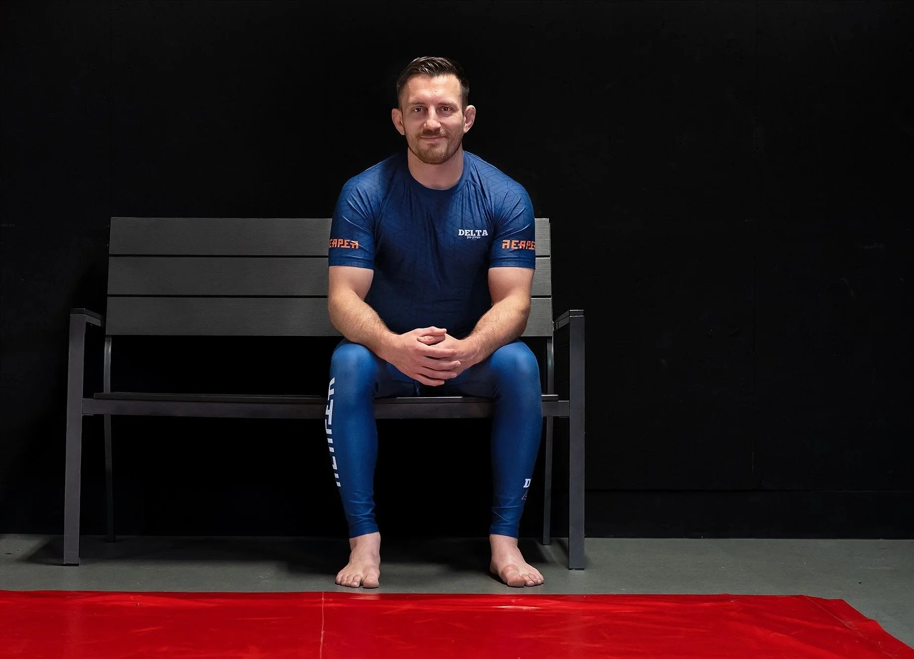 Man zit op een bankje in een No-Gi sportoutfit, met een zwarte achtergrond, op de rand van een rode mat.