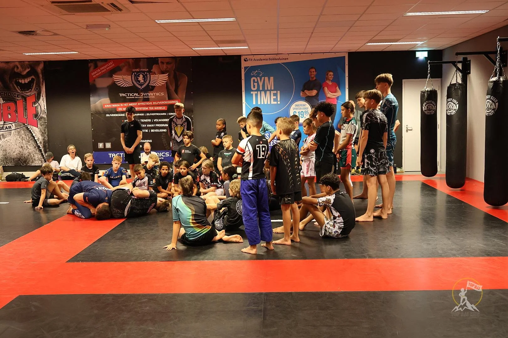 Groep kinderen en volwassenen in een sportschool, kinderen zitten op de grond terwijl een persoon op de grond ligt, mogelijk instructeur. Andere kinderen staan of zitten rondom de groep, een jongen in een blauwe broek staat in de rechter helft. Op de achtergrond posters en zogeheten bokszakken hangend aan de muur.