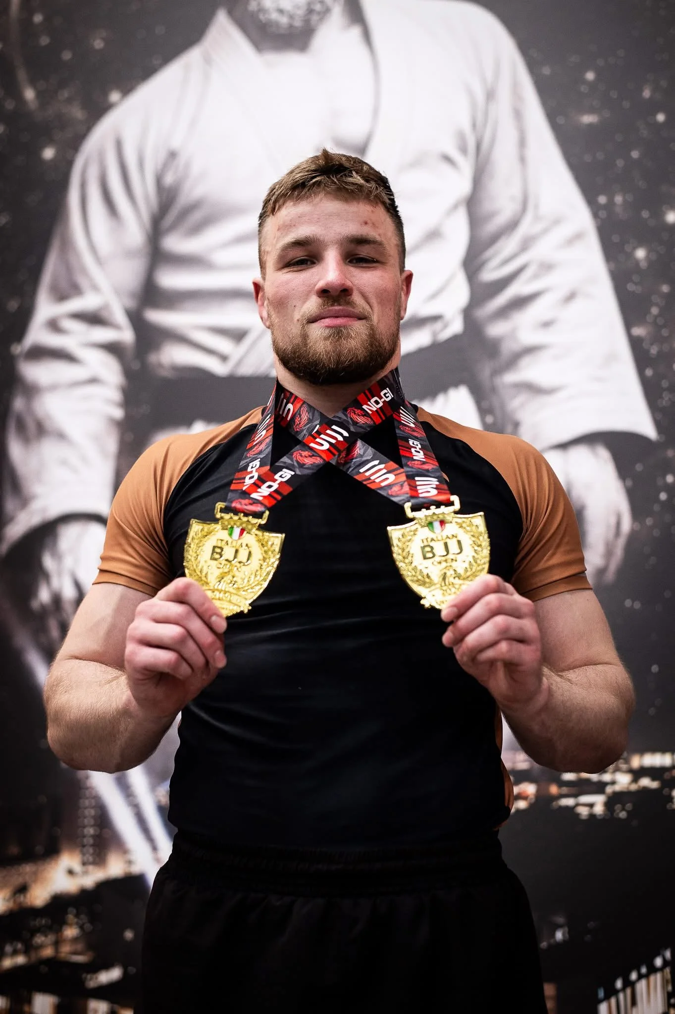 Een man met een baard en een korte coupe poseert trots met twee gouden medailles, tegen een achtergrond die lijkt op het maanoppervlak.