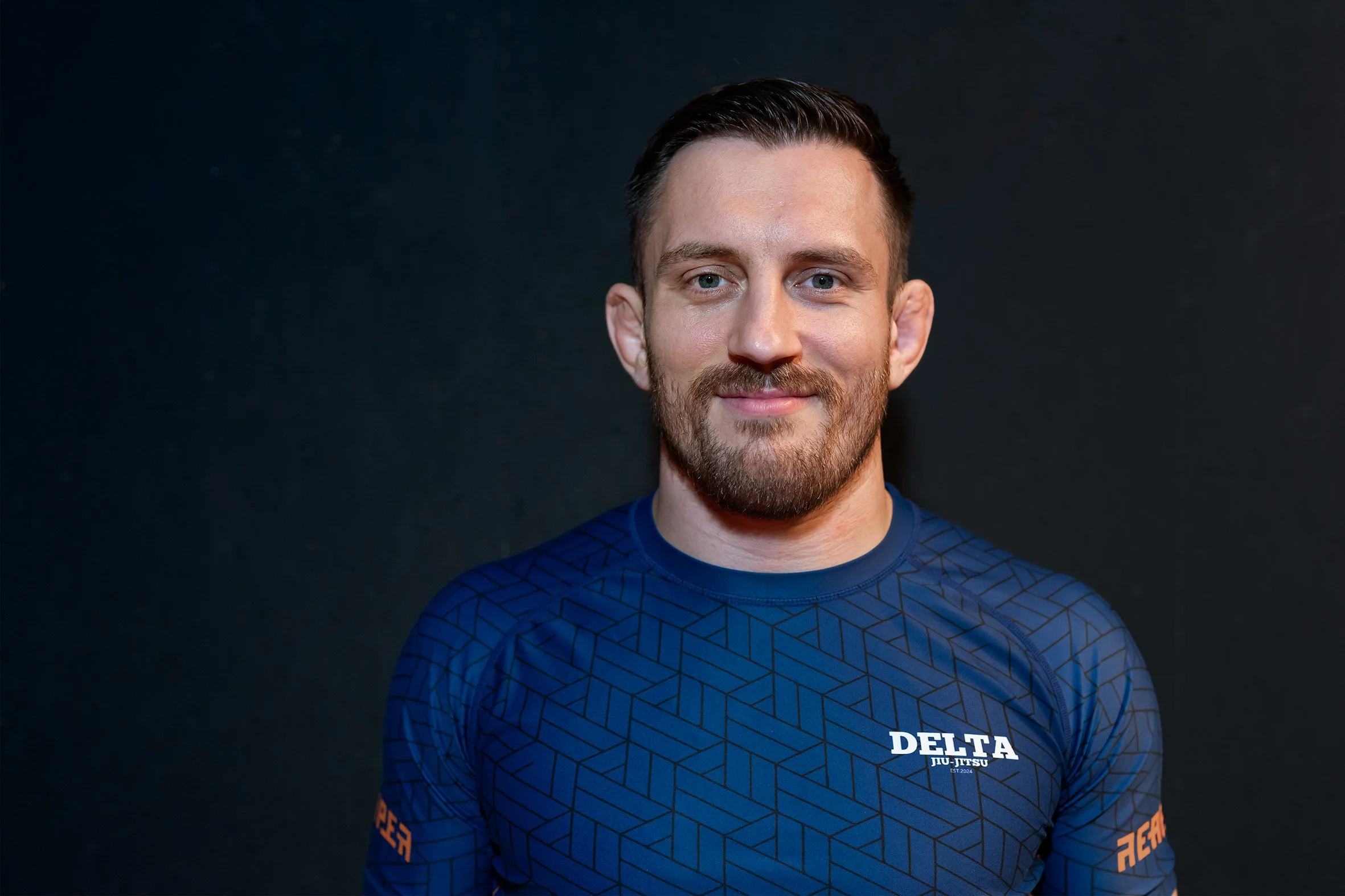 Man met een korte baard en zwarte haar, glimlacht, draagt een blauw sportshirt met tekst 'DELTA JU-JITSU, EST. 2024' op de voorkant, staart voor een donkere achtergrond.