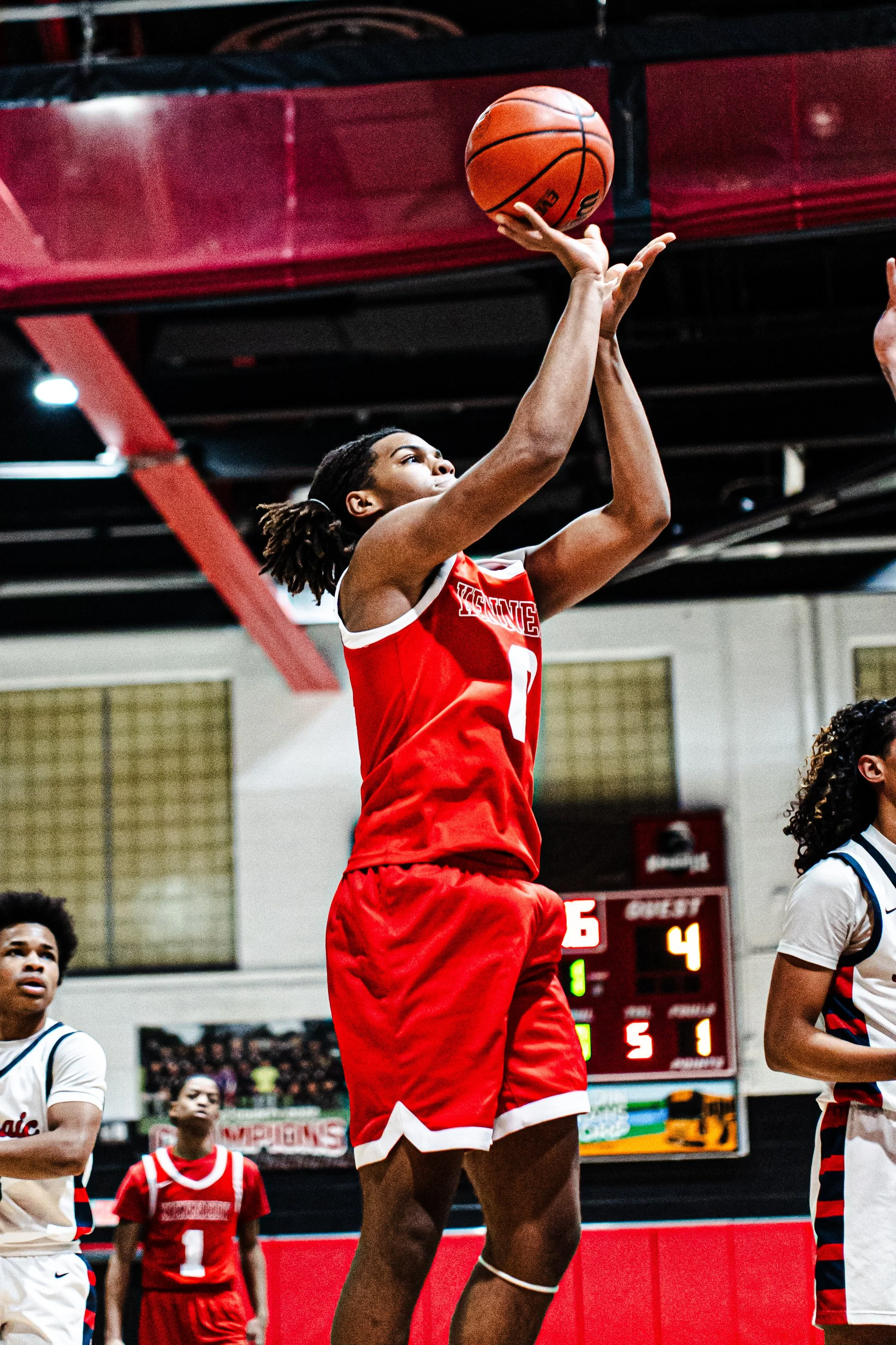 A female basketball player in red jersey and shorts shooting a basketball during a game in an indoor gym, with other players and a digital scoreboard visible in the background.