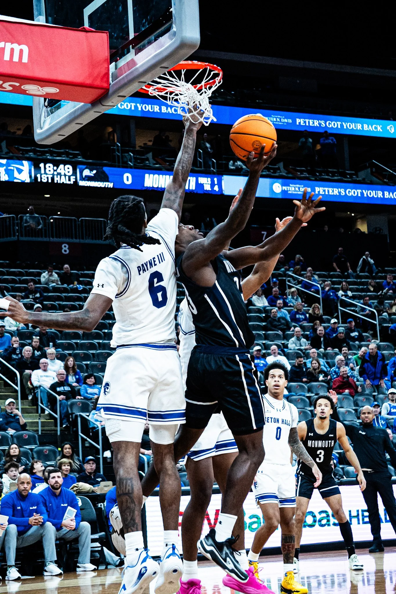 Two basketball players are jumping to reach the ball near the basket, with one in a white jersey and the other in a black jersey. The crowd in the background watches the game.