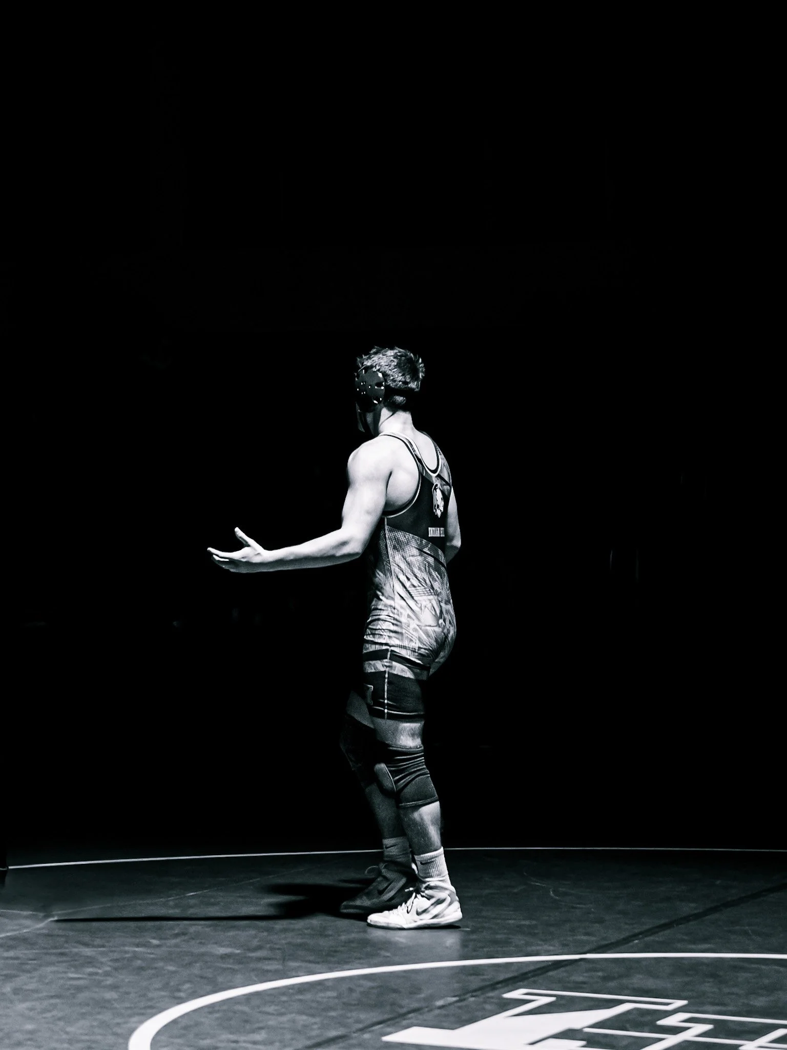 A basketball player in a tank top and shorts standing on a basketball court, viewed from behind. The scene is in black and white with a dark background.