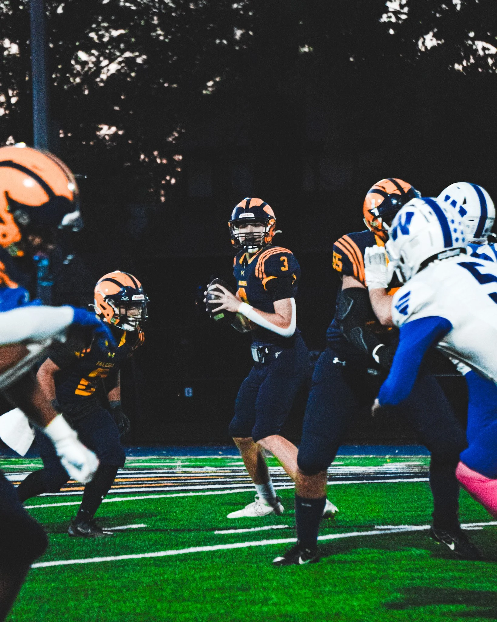 A football game is taking place at night on a field with bright green turf and colorful yard lines. Players from two teams, one in black and orange uniforms and the other in white and blue uniforms, are actively engaged in the game. The focus is on a