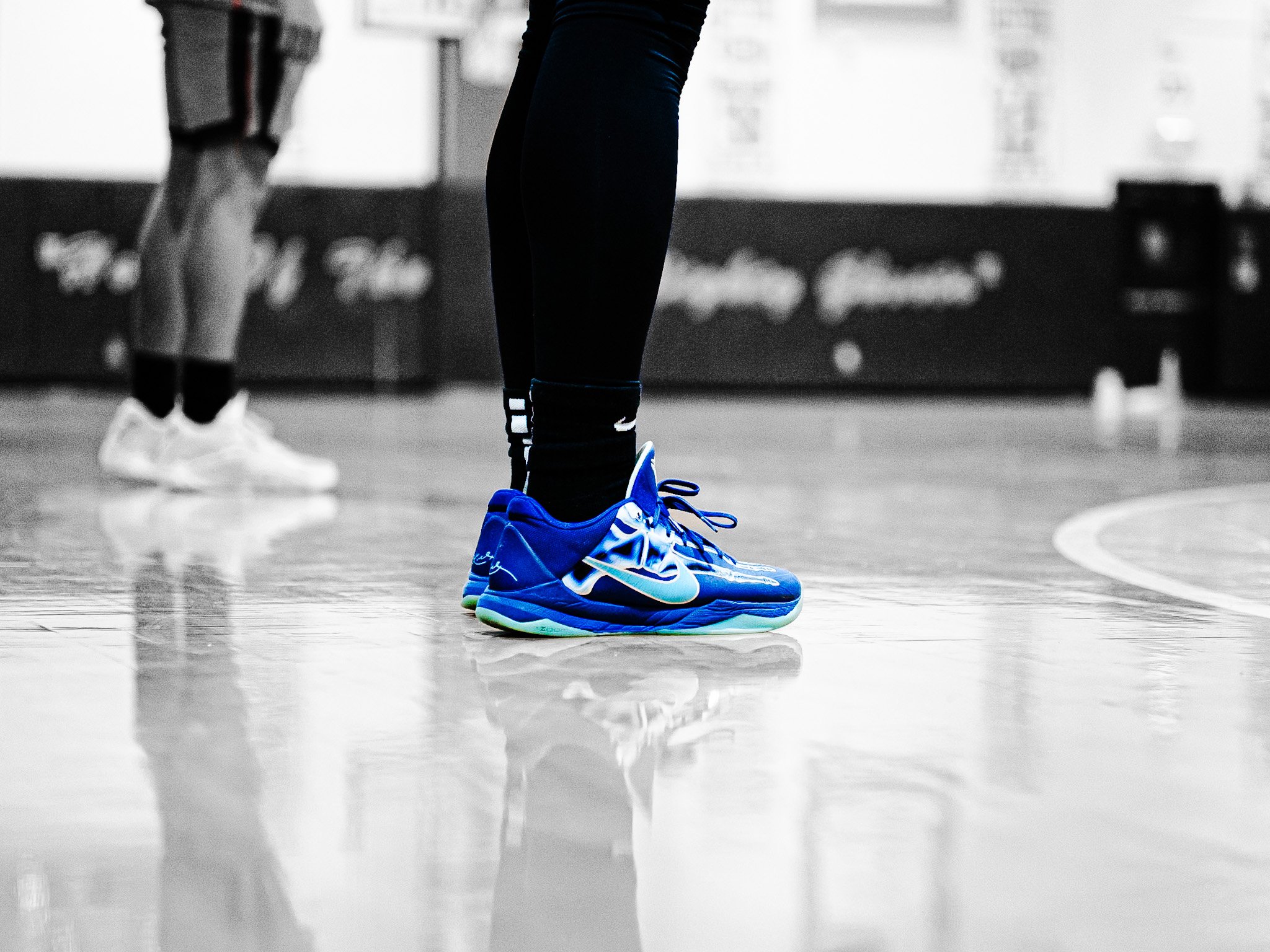 Close-up of two people standing on a gym floor, focusing on their legs and feet. One person wears bright blue athletic shoes and black leggings, while the other wears white shoes and shorts.
