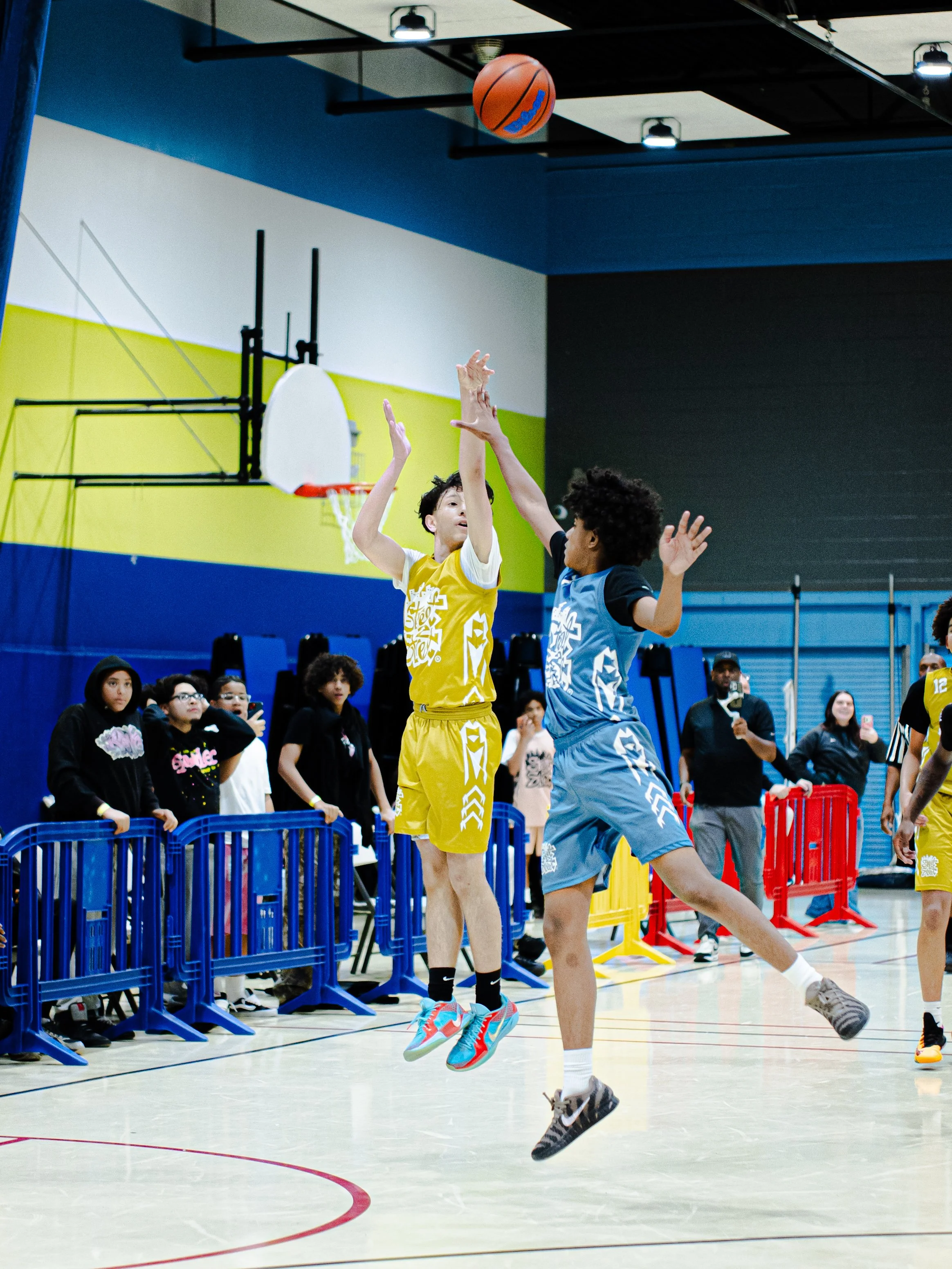 Two young basketball players, one in a yellow jersey and the other in a light blue jersey, jump in the air competing for the ball during a game in an indoor gymnasium. Spectators watch from behind a blue barrier along the sidelines.