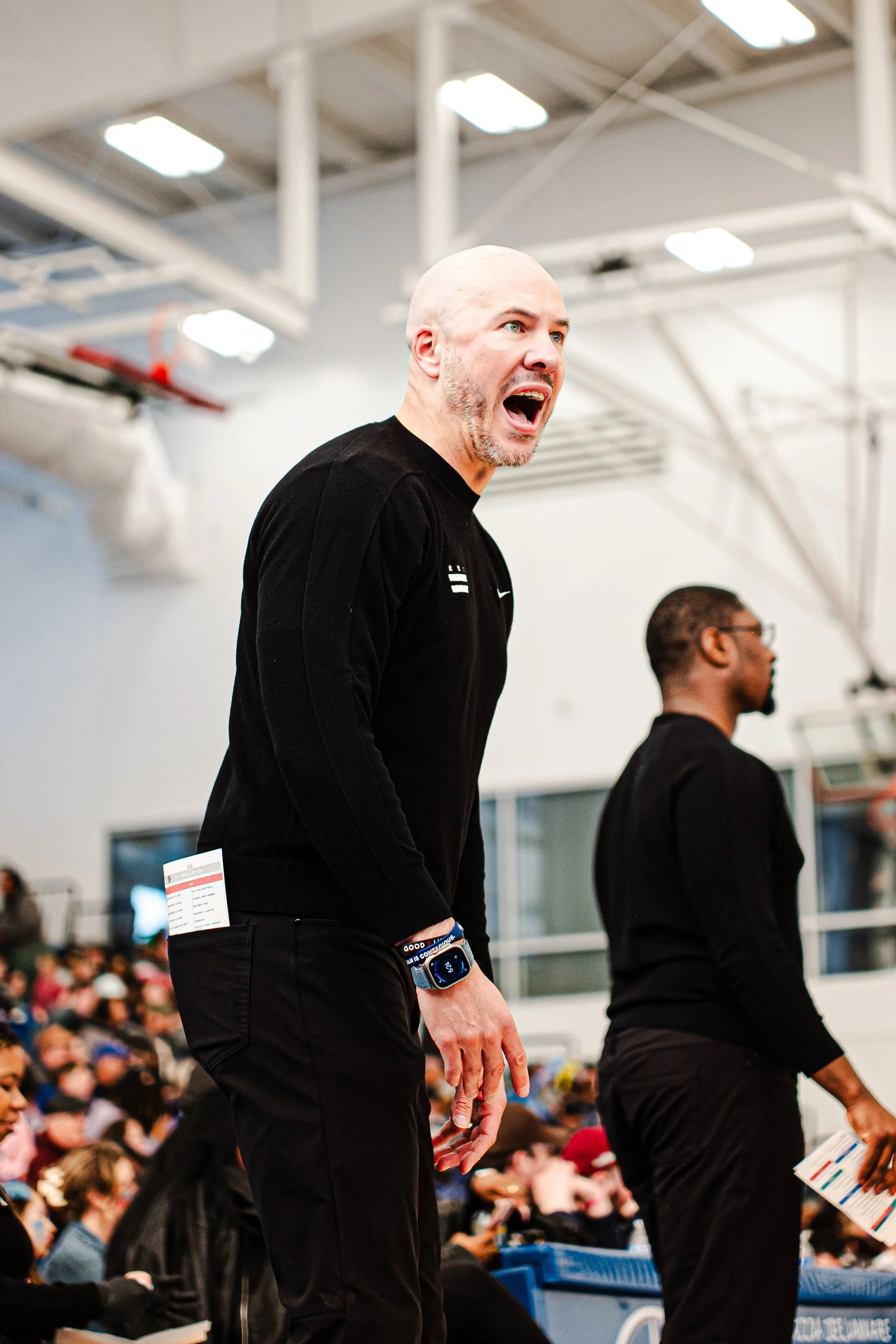 A man with a shaved head and beard wearing a black long sleeve shirt and a sports watch, standing among a crowd in an indoor arena. The crowd is seated and appears to be attending an event.
