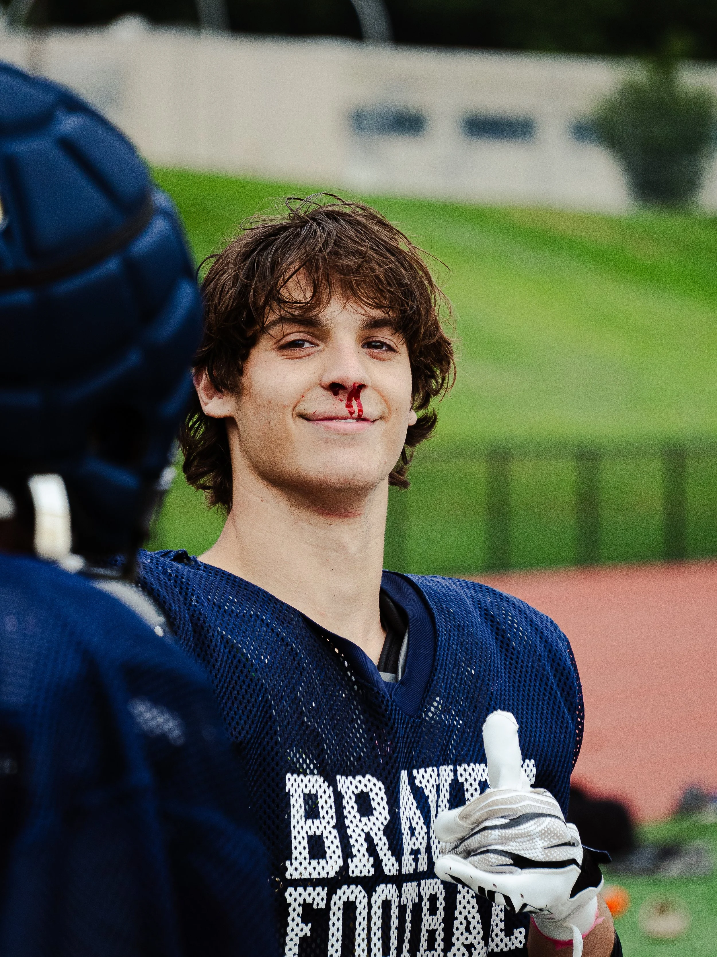 A young male football player with a nosebleed smiling, wearing a dark blue jersey and sports gloves, standing on a football field.