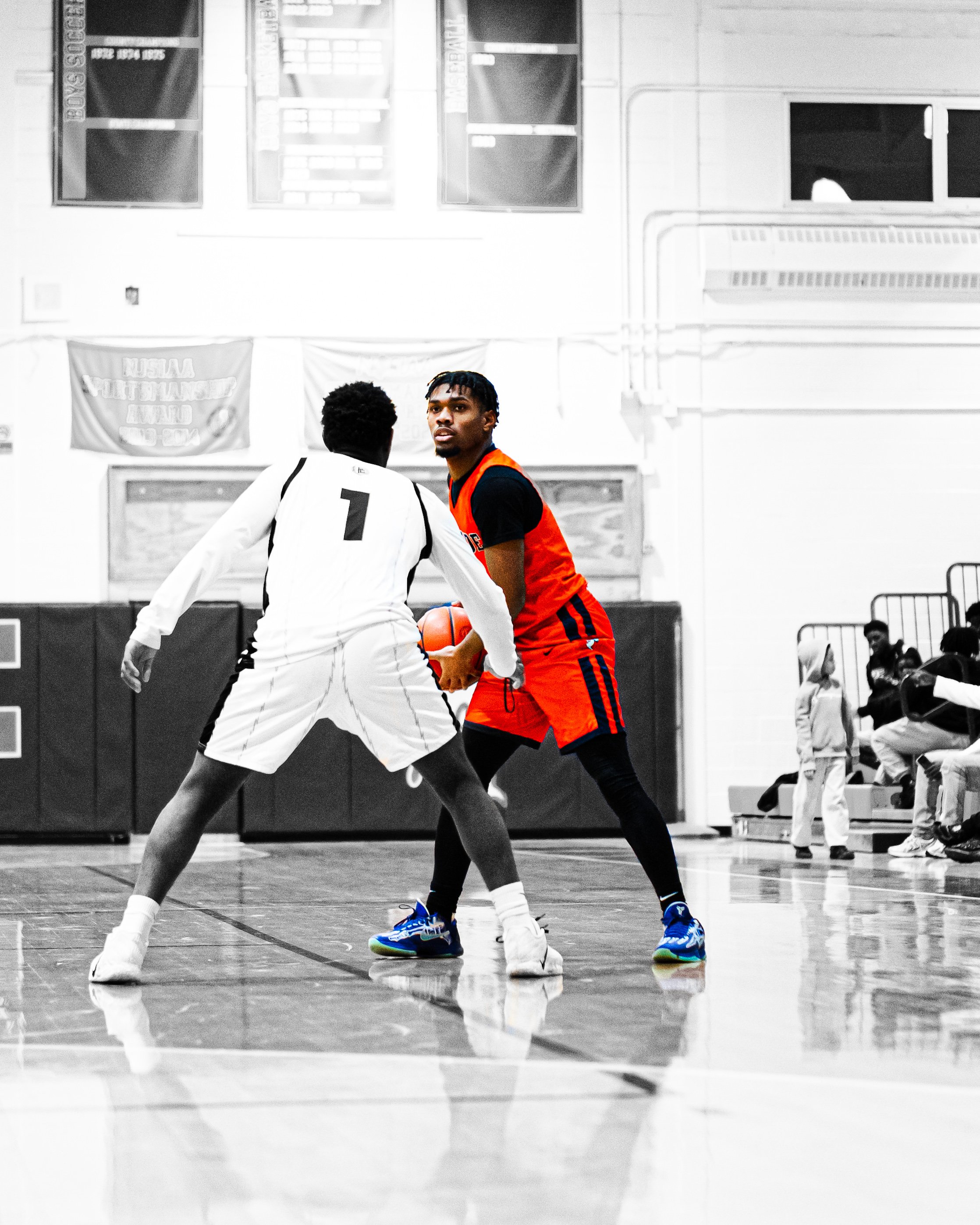 Two basketball players are playing a game in a gymnasium. One player is wearing a white uniform with the number 1, and the other player is wearing a red and black uniform. They are holding a basketball and appear to be contesting a play. The gym has 