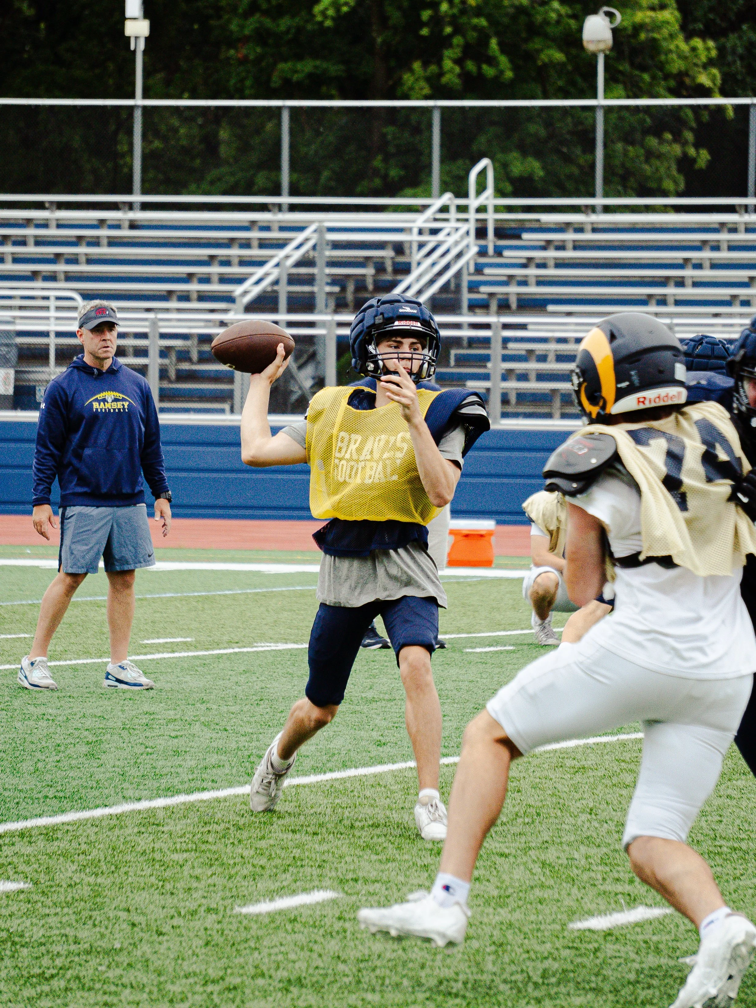 A youth football player wearing a helmet and yellow jersey throwing a football during practice on a turf field, with a coach and other players visible in the background.