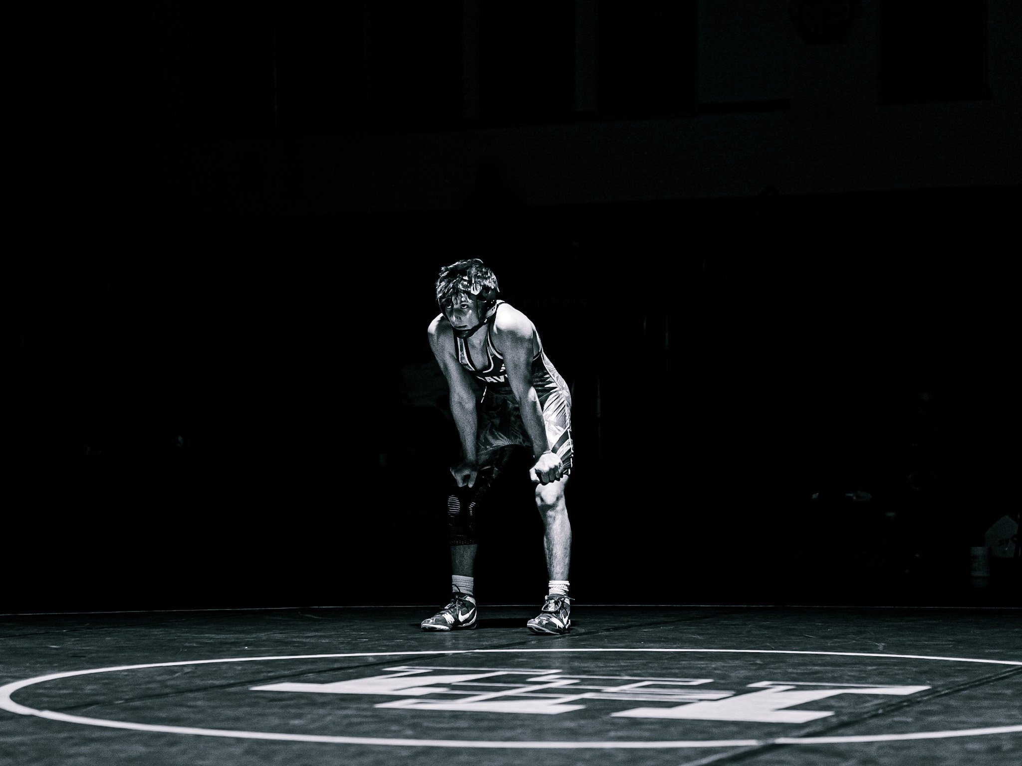 A person dressed in sportswear, including a singlet and shorts, is crouching on a wrestling mat, preparing for a match in a dark room.