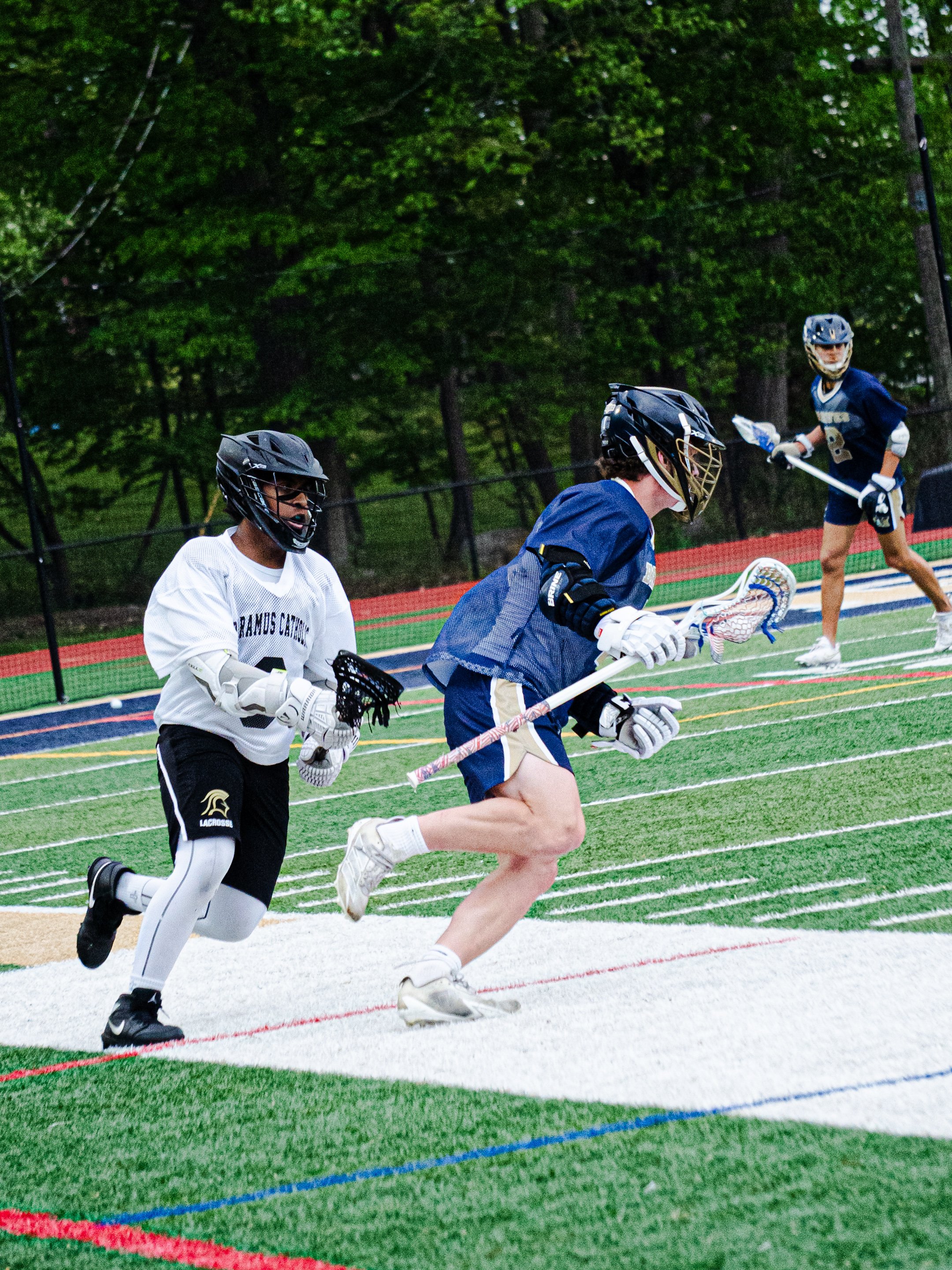 Three young boys playing lacrosse on a field with trees in the background. Two are running and one is standing, all wearing helmets, gloves, and sports uniforms.