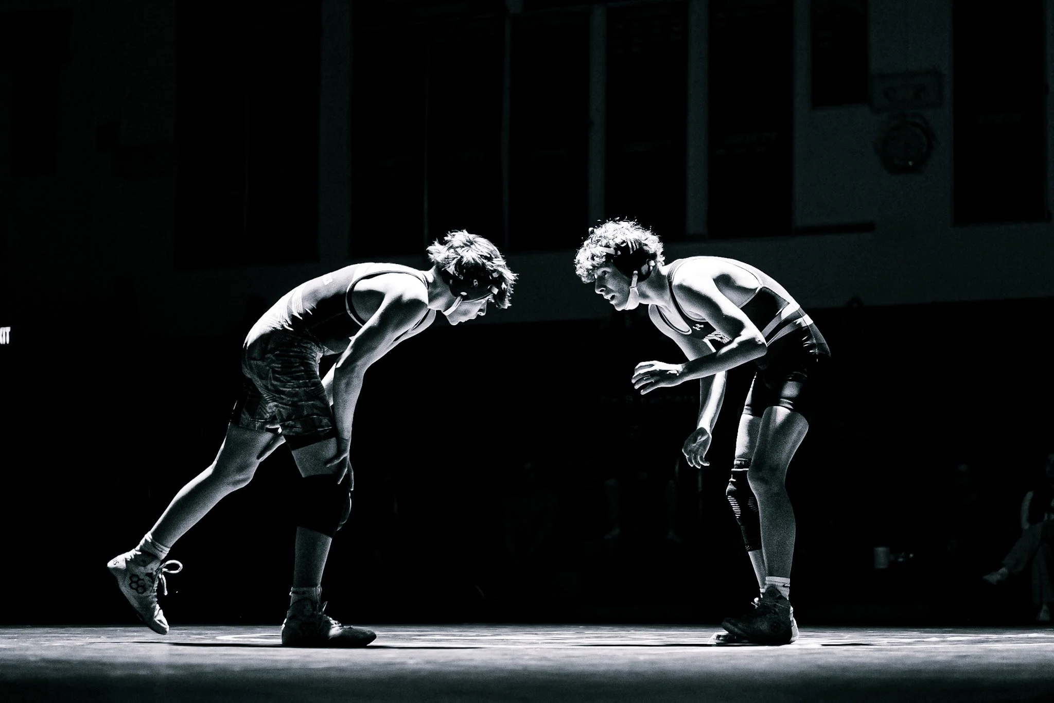 Two wrestlers in singlets and protective headgear bent over facing each other on a wrestling mat in a dark gym or arena, with a referee or coach partially visible in the background.