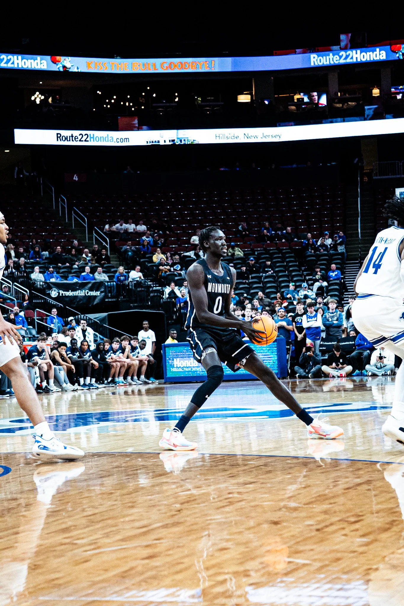A basketball game in an indoor arena, with a player in a black jersey holding the ball and preparing to make a move. Other players and spectators are visible in the background.