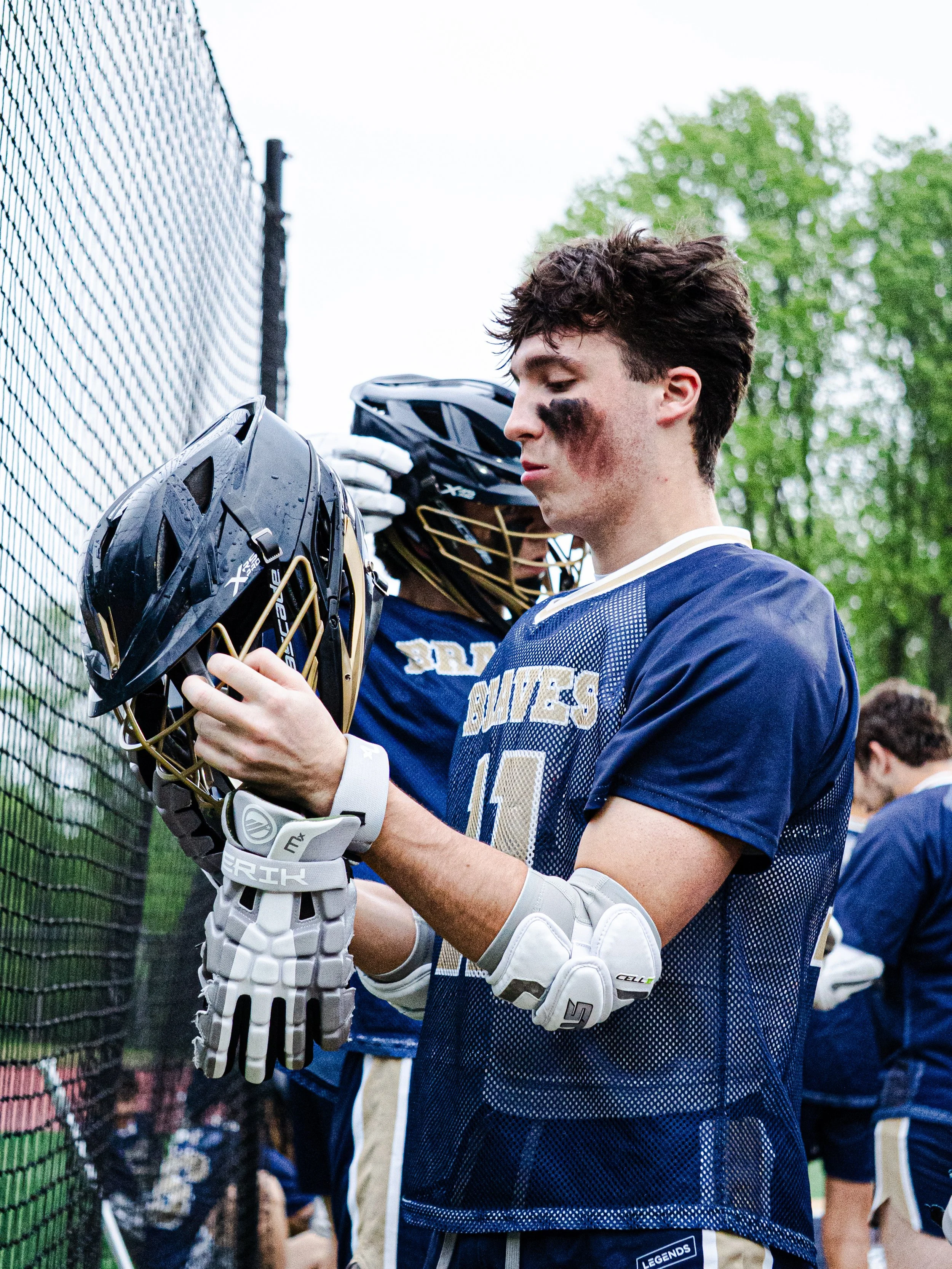 A young male lacrosse player with black eye black on his face, wearing a navy blue team jersey with 'RIVALS' on it and holding a lacrosse helmet, standing near a chain-link fence during a game or practice.