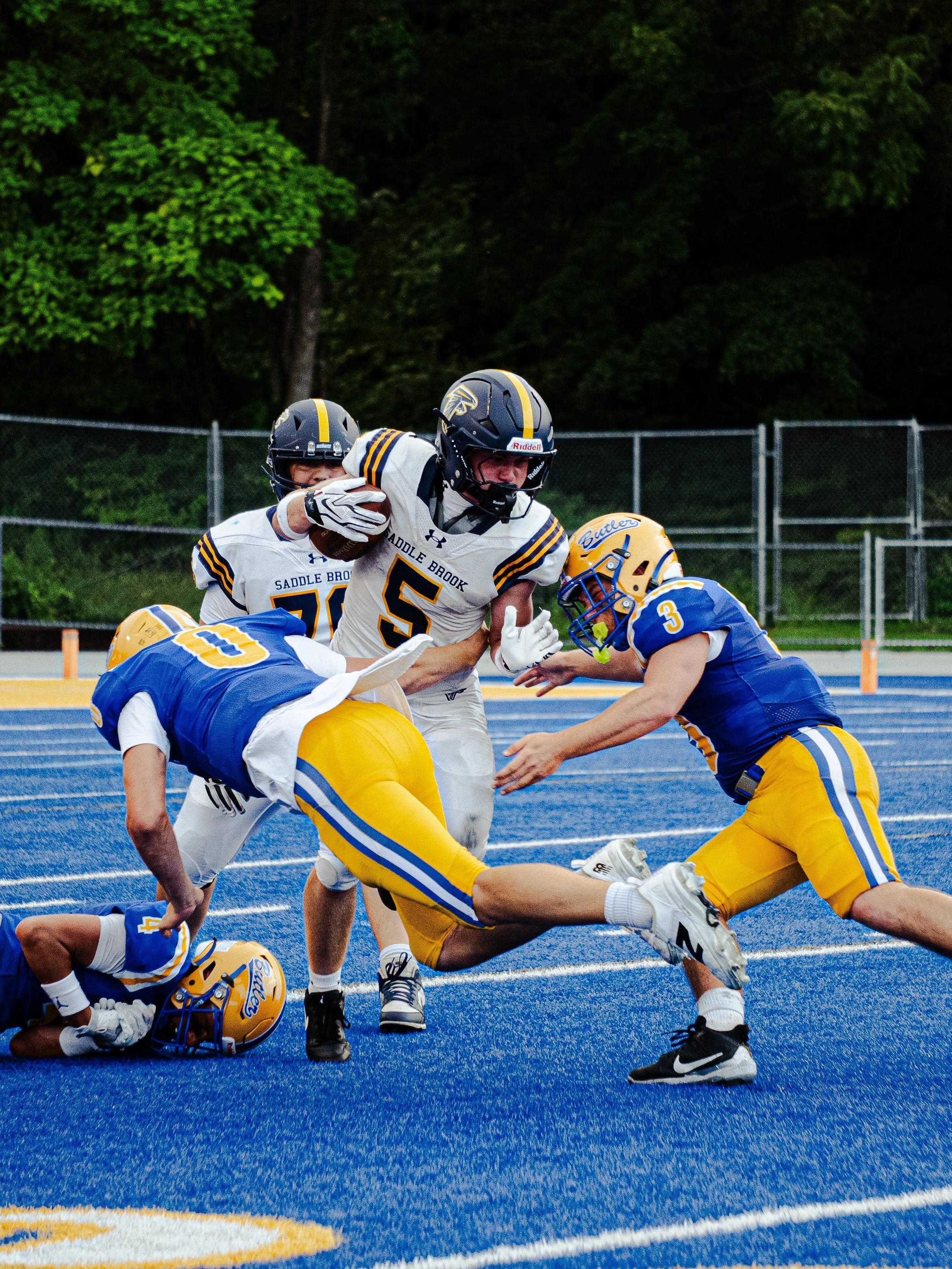High school football game with players on a blue turf field, one in a white jersey with the number 55, and the others in blue and yellow uniforms. Players are engaged in a tackle play with one player on the ground and others reaching for the ball.